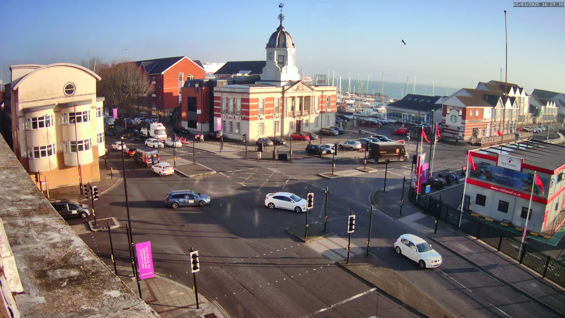 A high-angle view of a town square with a clock tower and several buildings surrounding a multi-way intersection with cars and traffic lights under a clear, sunny sky.