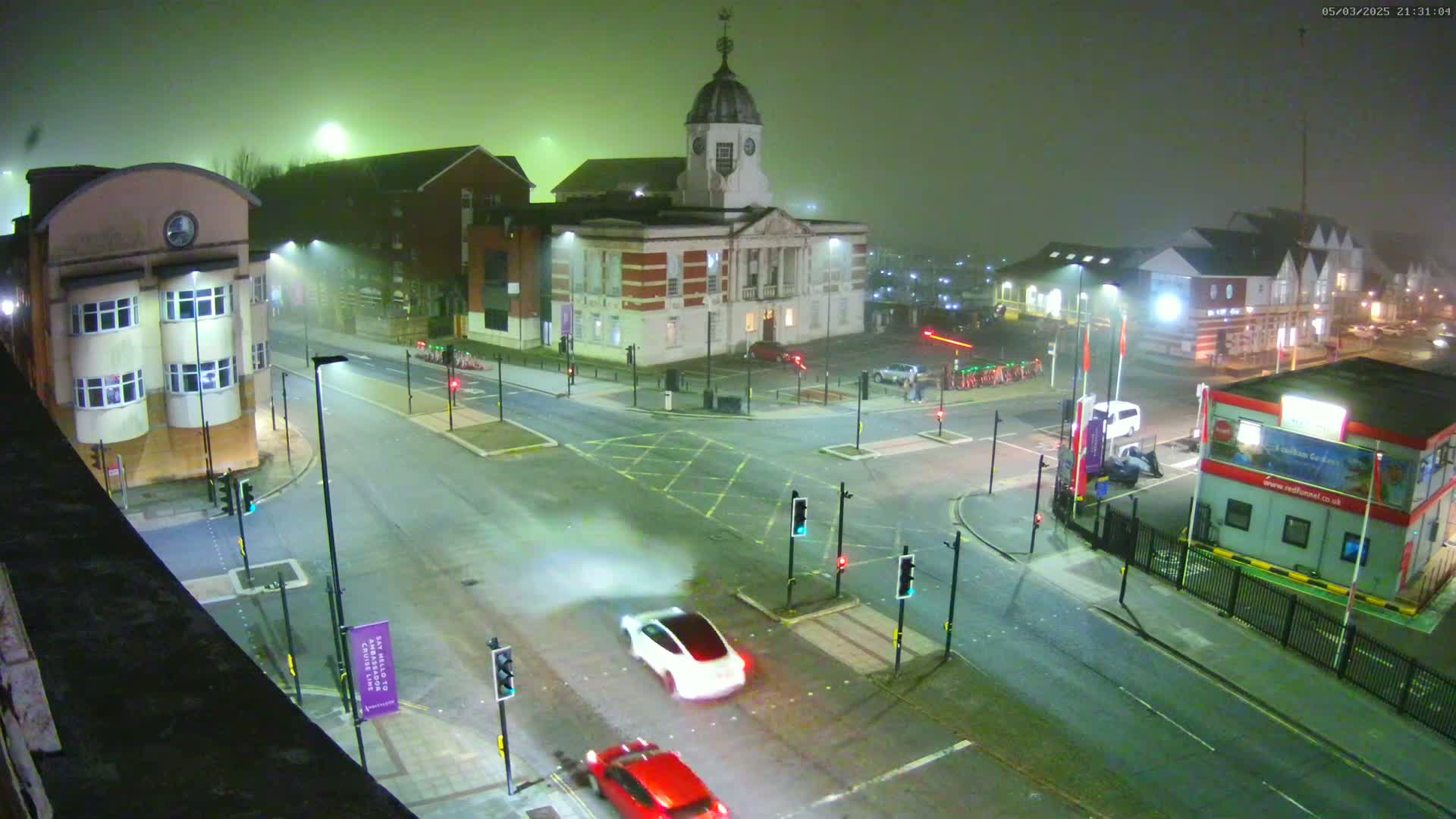 A foggy night scene shows a multi-lane intersection with several buildings, including a large brick building with a dome, and a few cars navigating the roadways under streetlights.