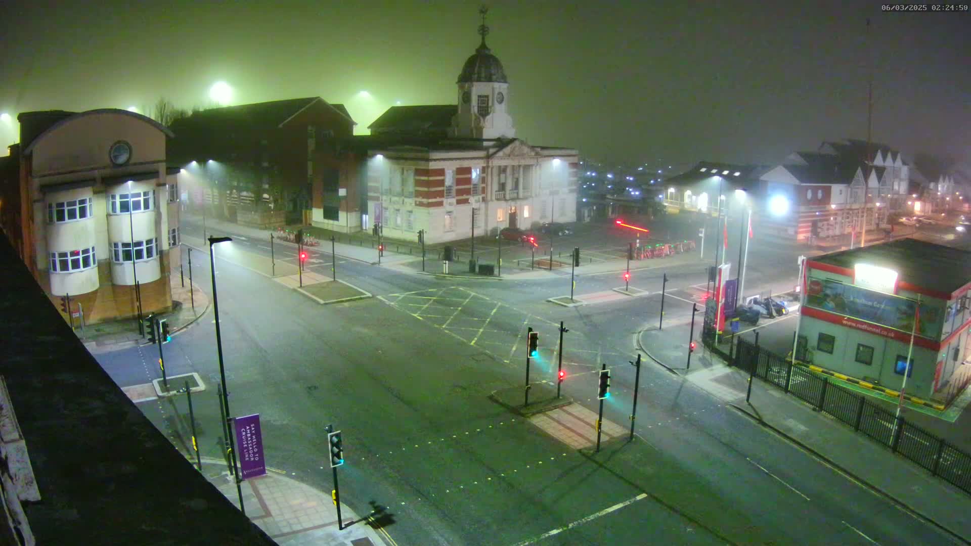 A foggy night scene shows a multi-way intersection with traffic lights, several buildings, and a few parked cars.