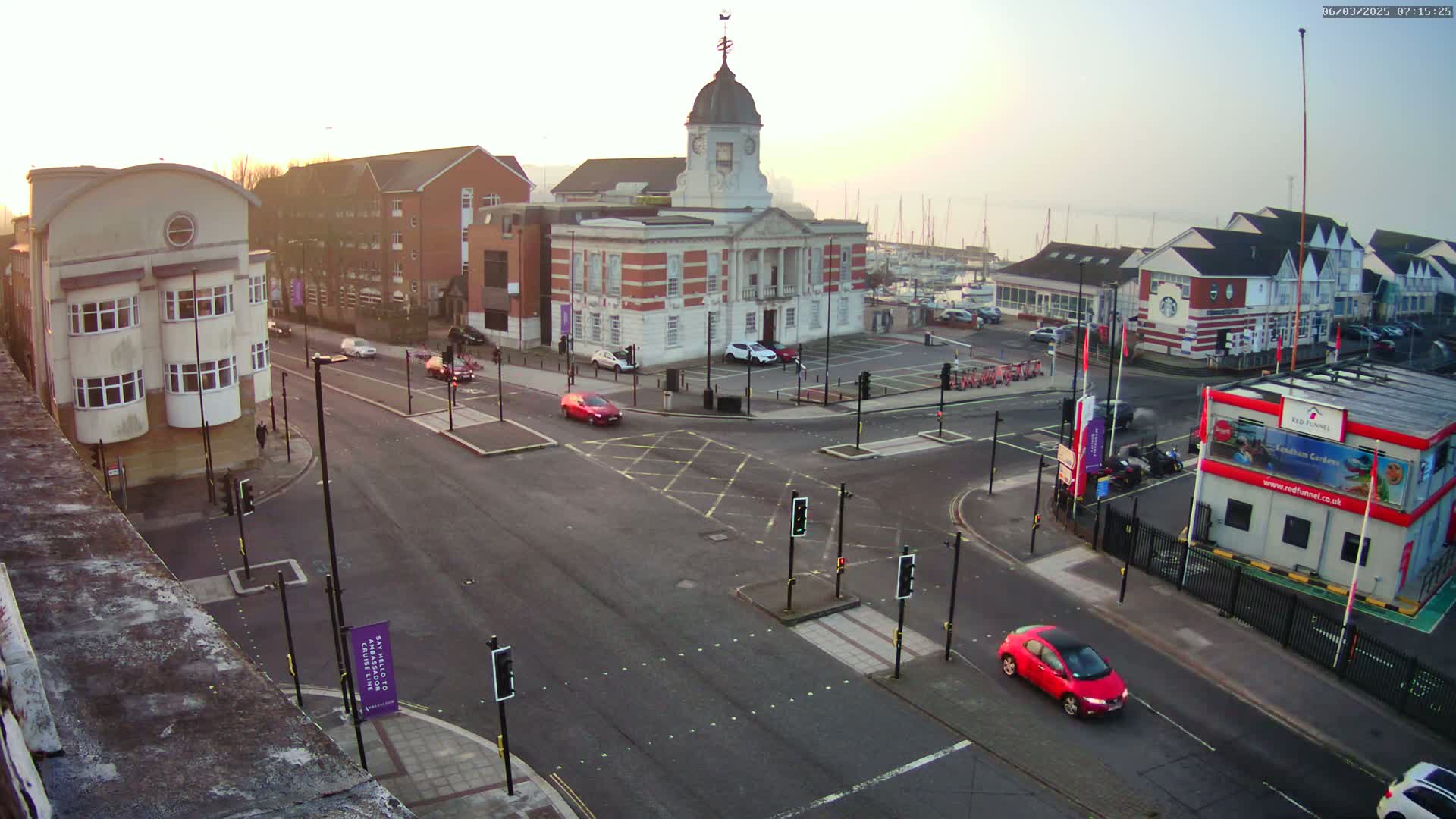 A bright, hazy sunrise illuminates a town square with a large brick building, several smaller buildings, a harbor with boats, and cars at a multi-way intersection.