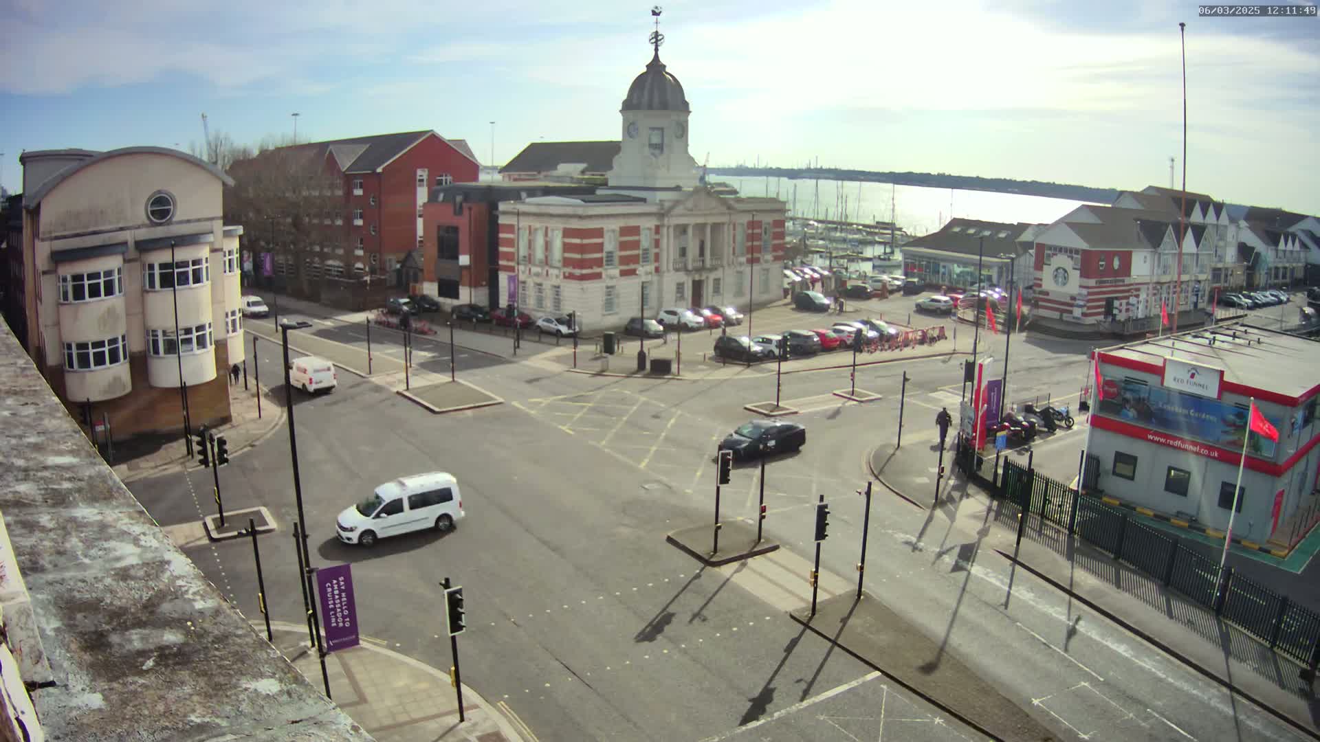 A sunny day reveals a town square with a brick building and a domed tower, surrounded by streets, cars, and a marina visible in the background.