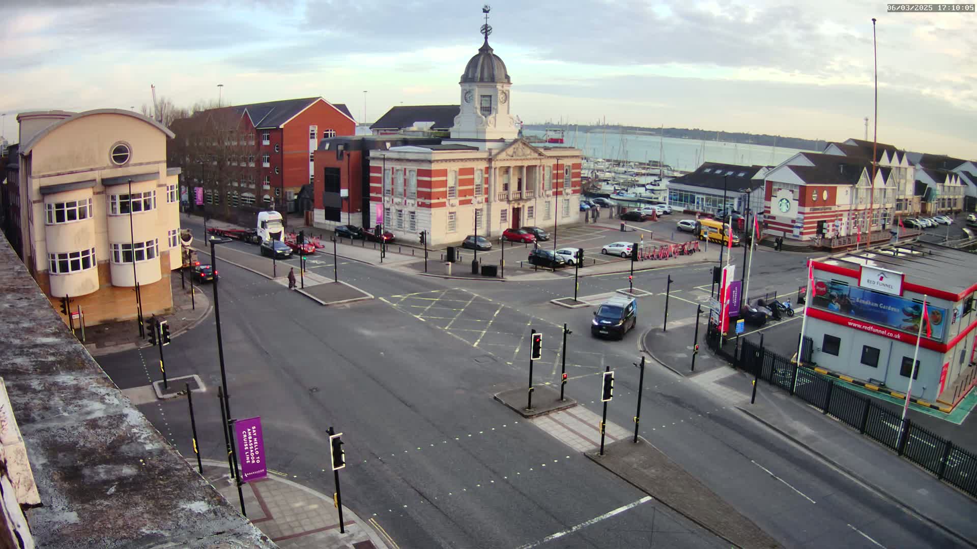 A street intersection with a brick building and clock tower in the center, surrounded by other buildings, cars, and a marina with boats in the background, under partly cloudy skies.