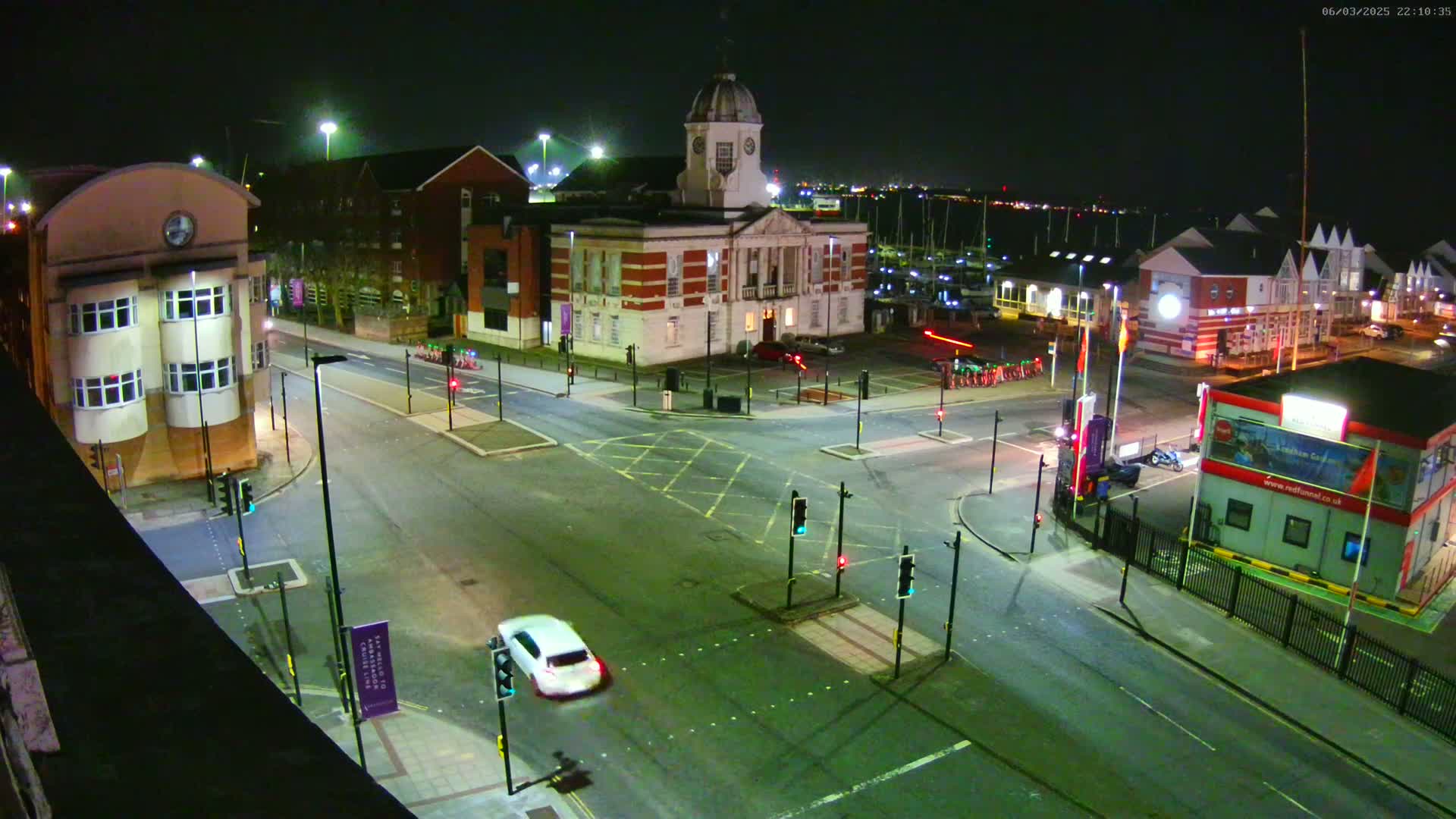 A nighttime aerial view of a city intersection with several buildings, including a large brick building with a dome, and a marina visible in the background under clear skies.