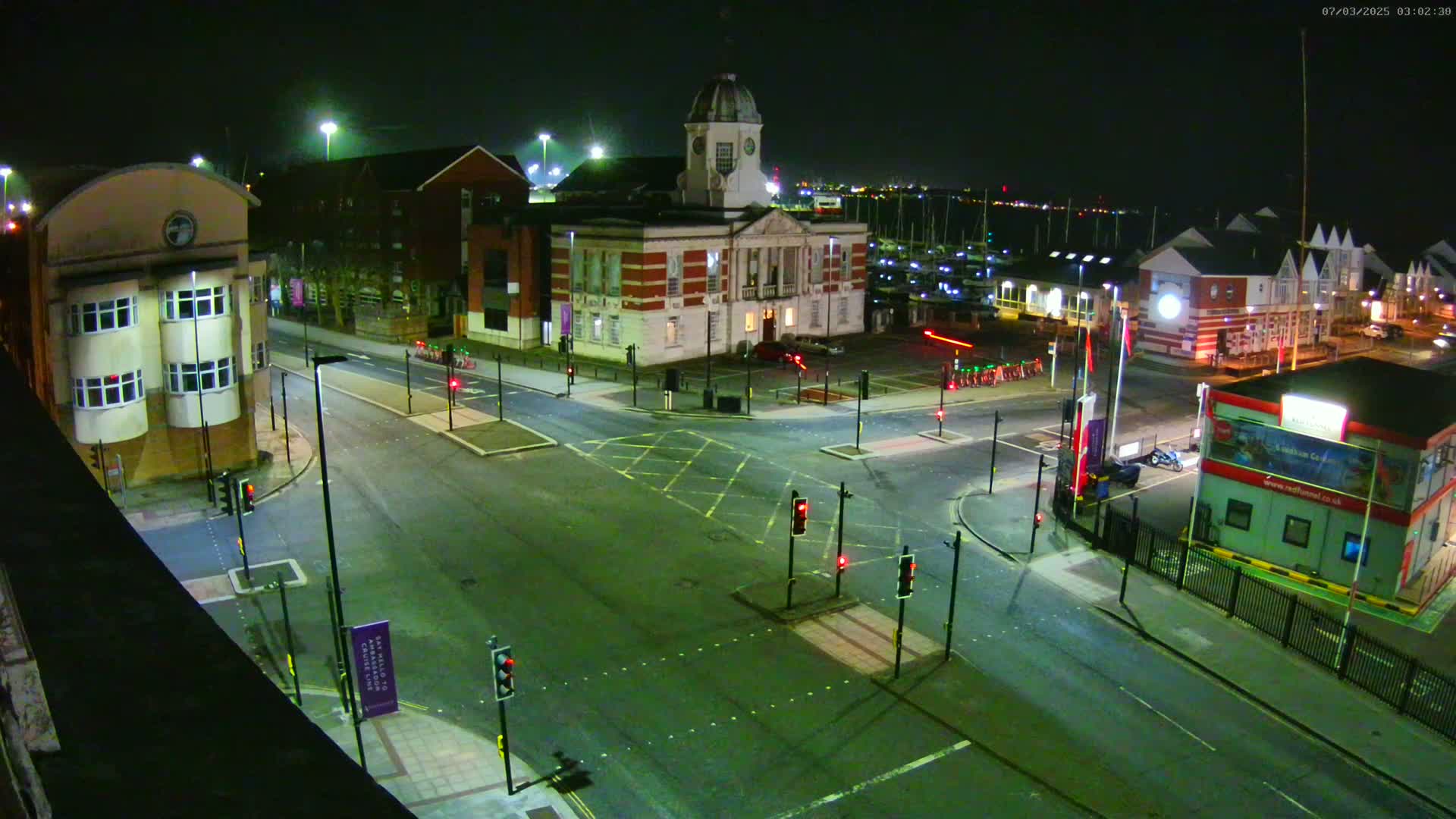 A nighttime, high-angle view of a street intersection in front of a brick building with a clock tower, near a marina with docked boats under clear skies.
