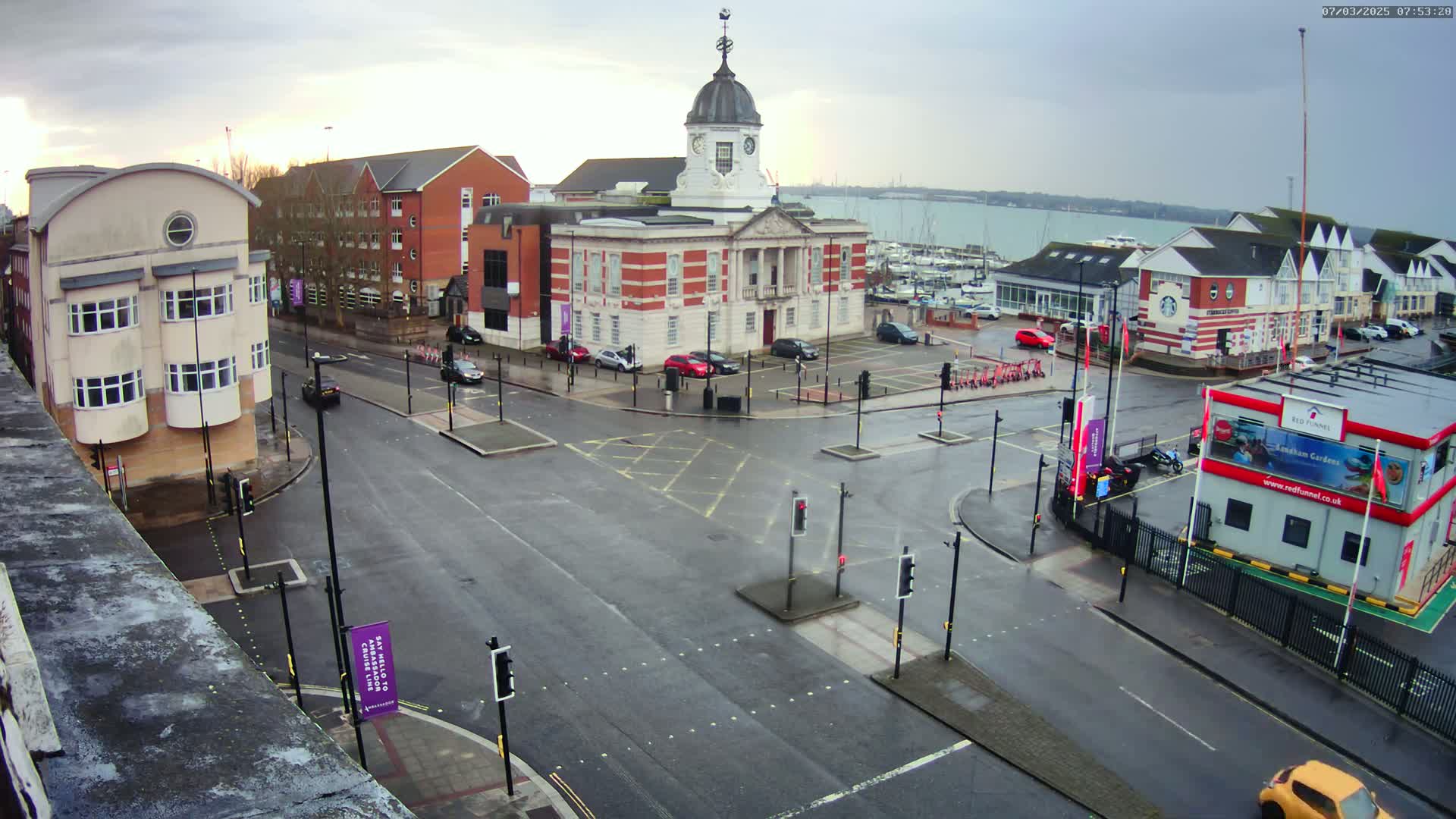 A wet, overcast day reveals a town square with a brick clock tower building at its center, surrounded by other buildings, roads, and a marina visible in the background.