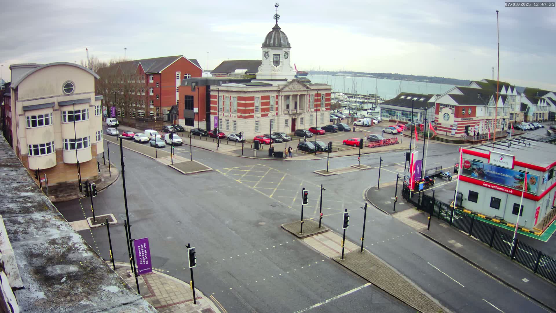 An overcast day reveals a town square with a brick building featuring a cupola, surrounded by other buildings, parked cars, and a harbor visible in the background.