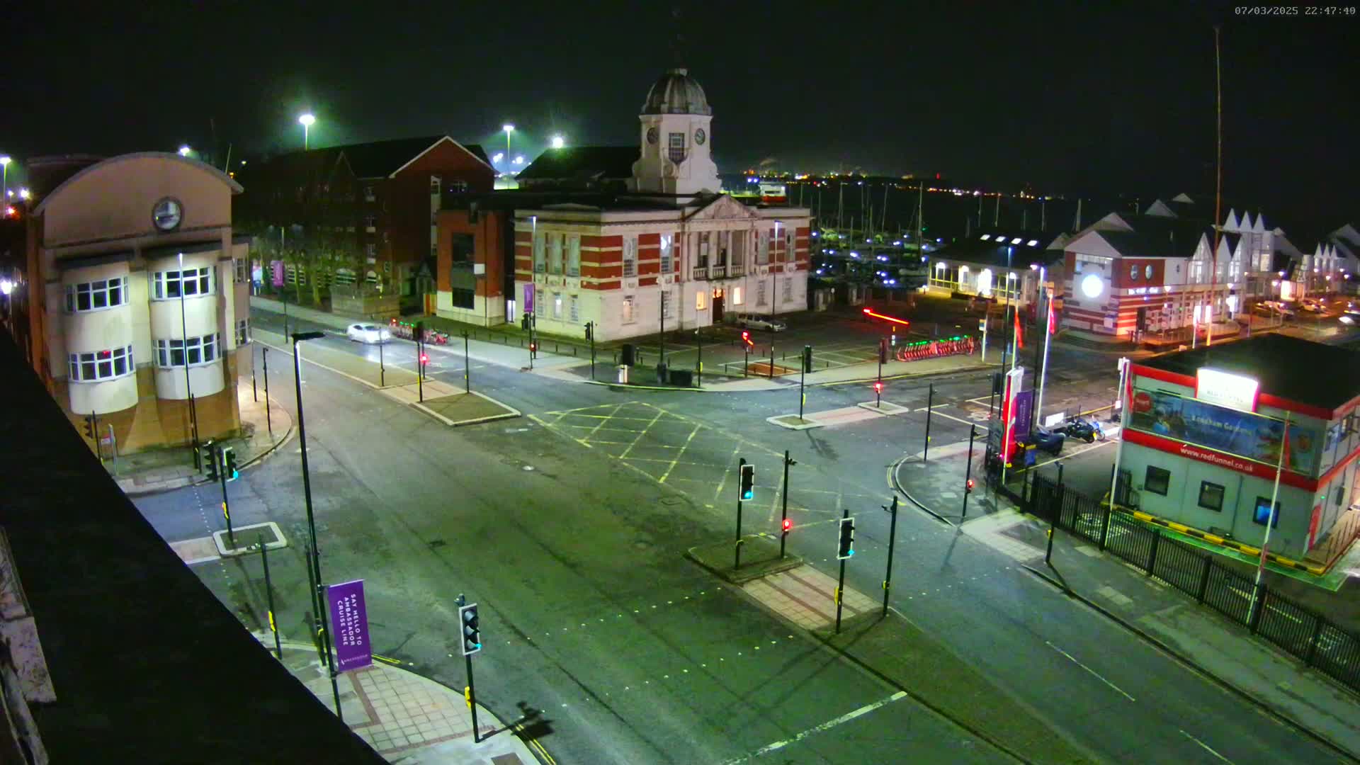 A nighttime aerial view of a town square with a large brick building at its center, surrounded by streets, traffic lights, and parked cars, under clear skies.