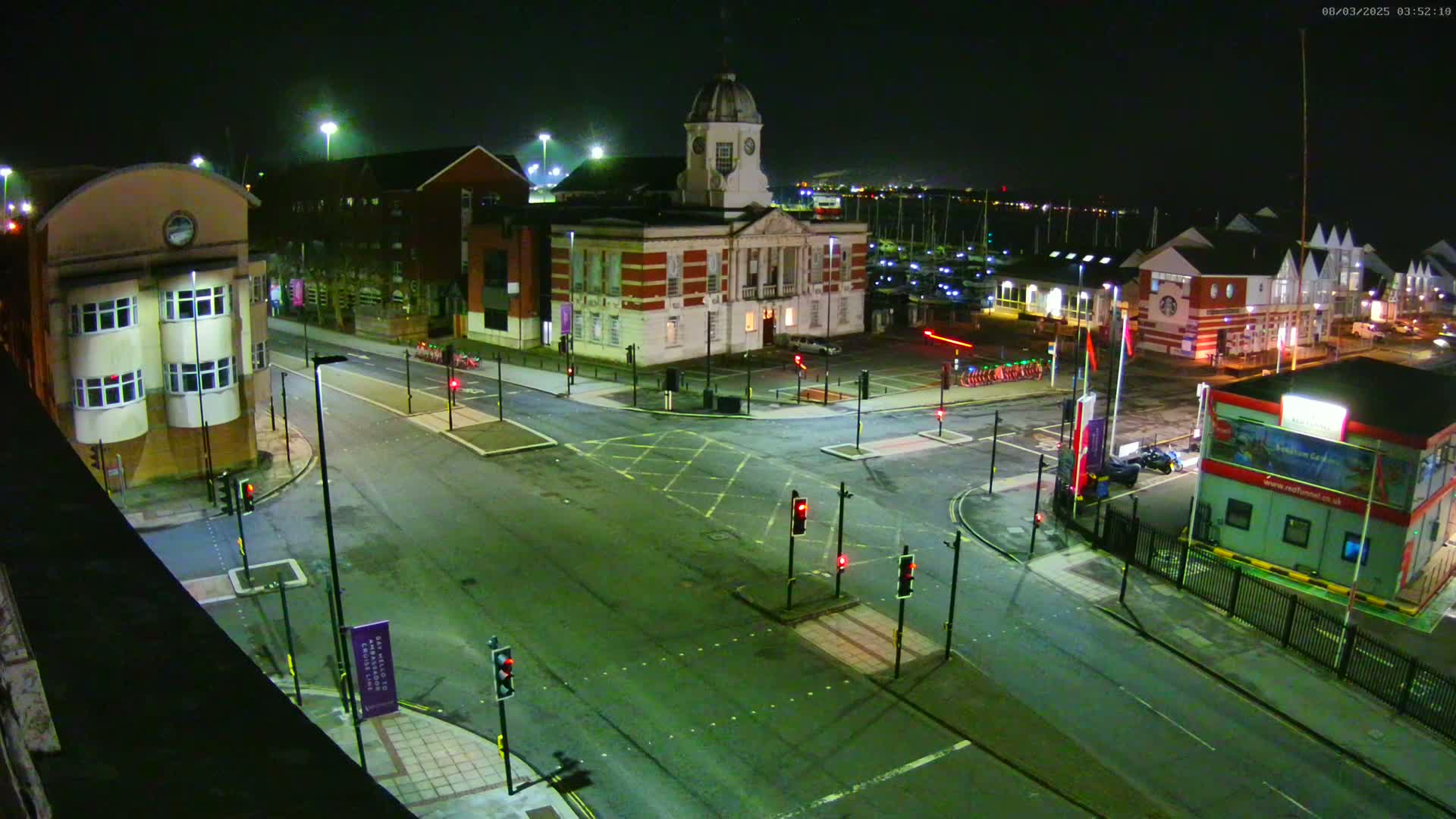 A nighttime high-angle view of an empty intersection with several buildings, including a large brick building with a dome, and a marina visible in the background.