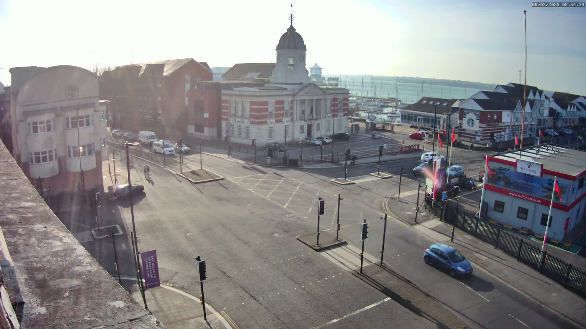A sunny day reveals a street intersection with several buildings, including a large brick building with a dome, and a marina with boats in the background.