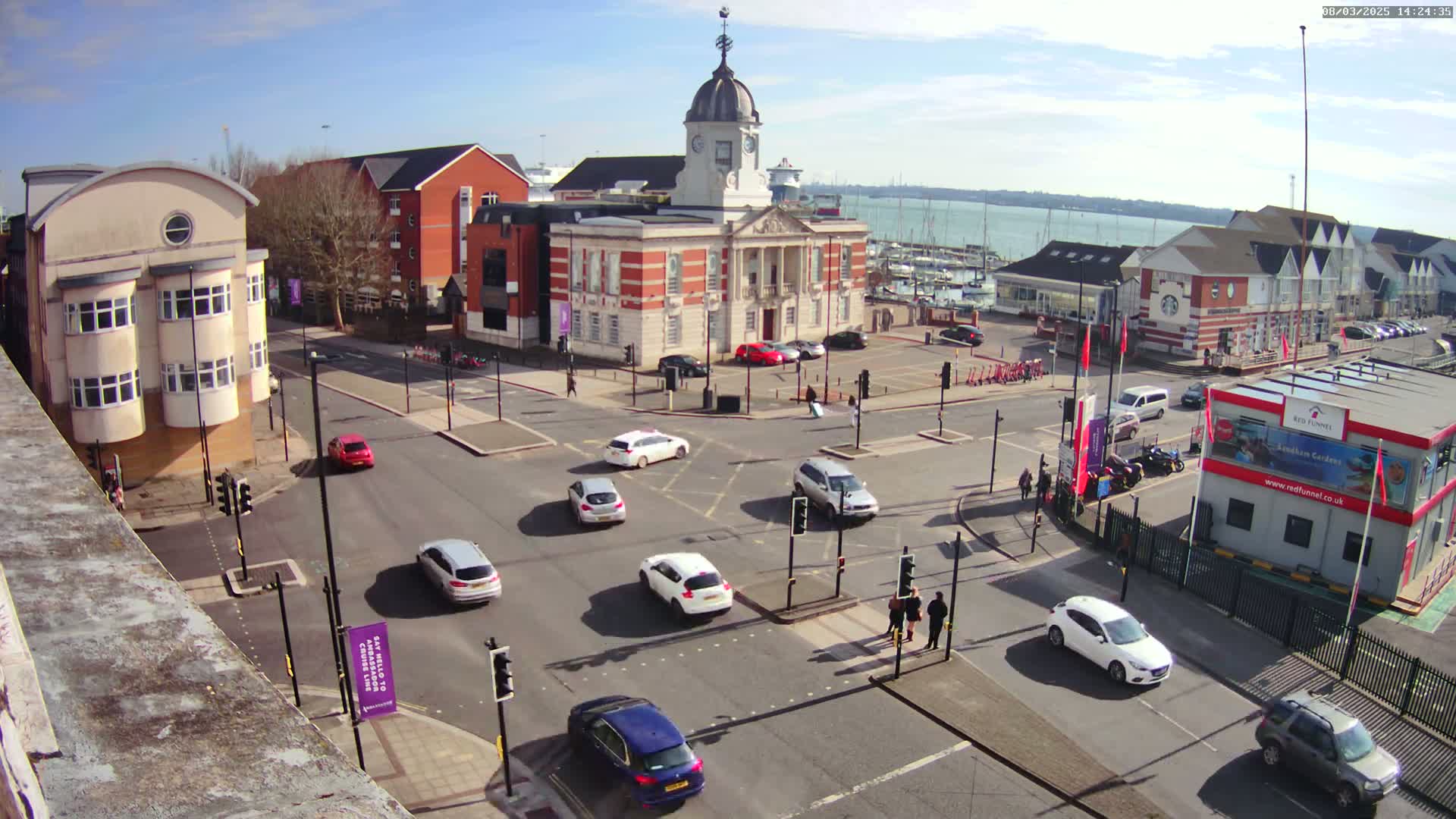 A sunny day shows a town square with a brick building at its center, surrounded by other buildings, several cars at a crossroads, and a body of water with boats in the distance.