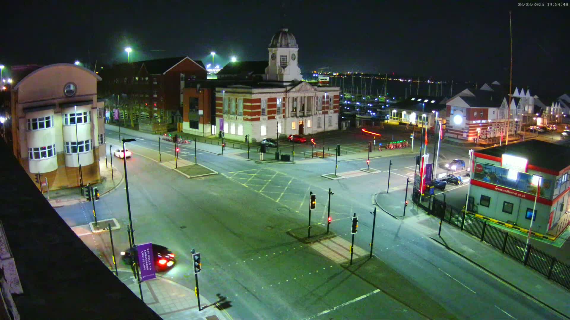 A nighttime, high-angle view of a street intersection near a marina with several buildings, including a large brick building with a dome, under clear skies.