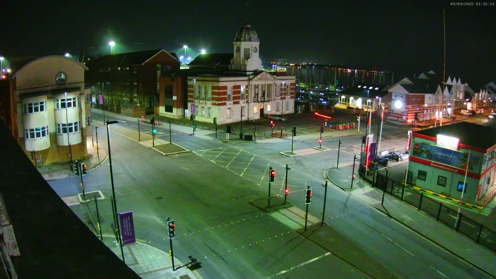 A nighttime aerial view of a town square with a large brick building, several smaller buildings, and a marina in the background, under clear skies.