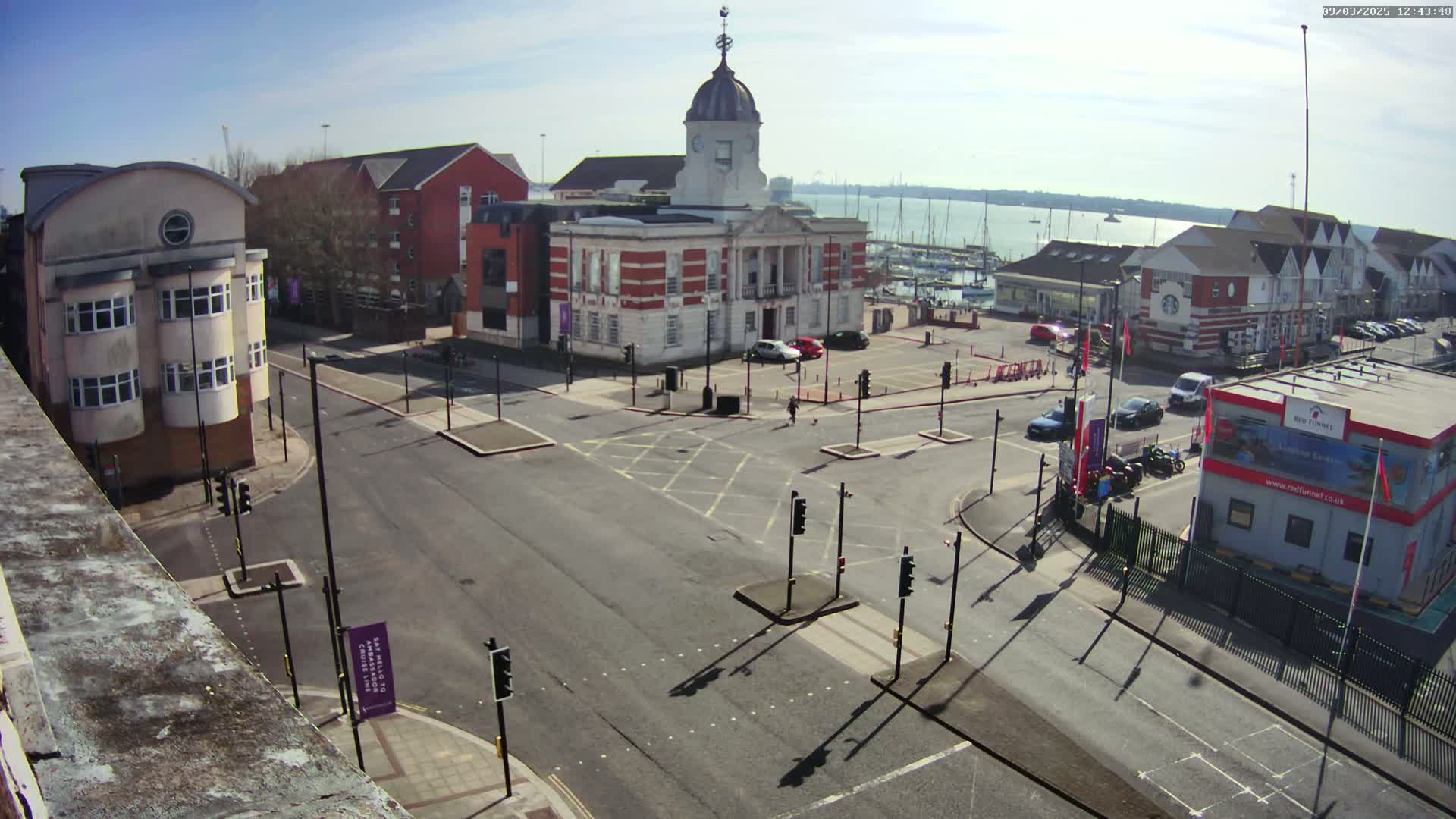 A sunny day reveals a town square with a brick building, surrounded by other structures and a marina visible in the background.