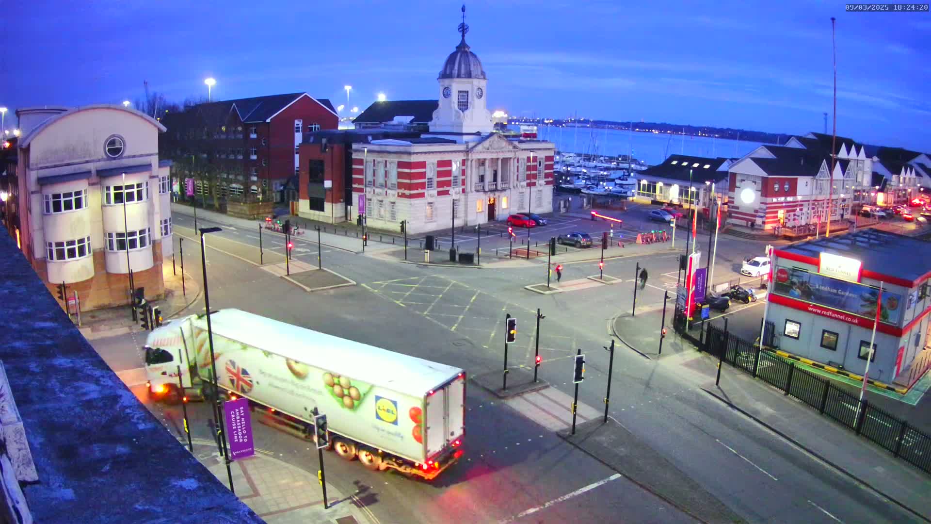 A large white truck drives through a town square at dusk near a harbor with many boats, passing a red brick building with a clock tower.
