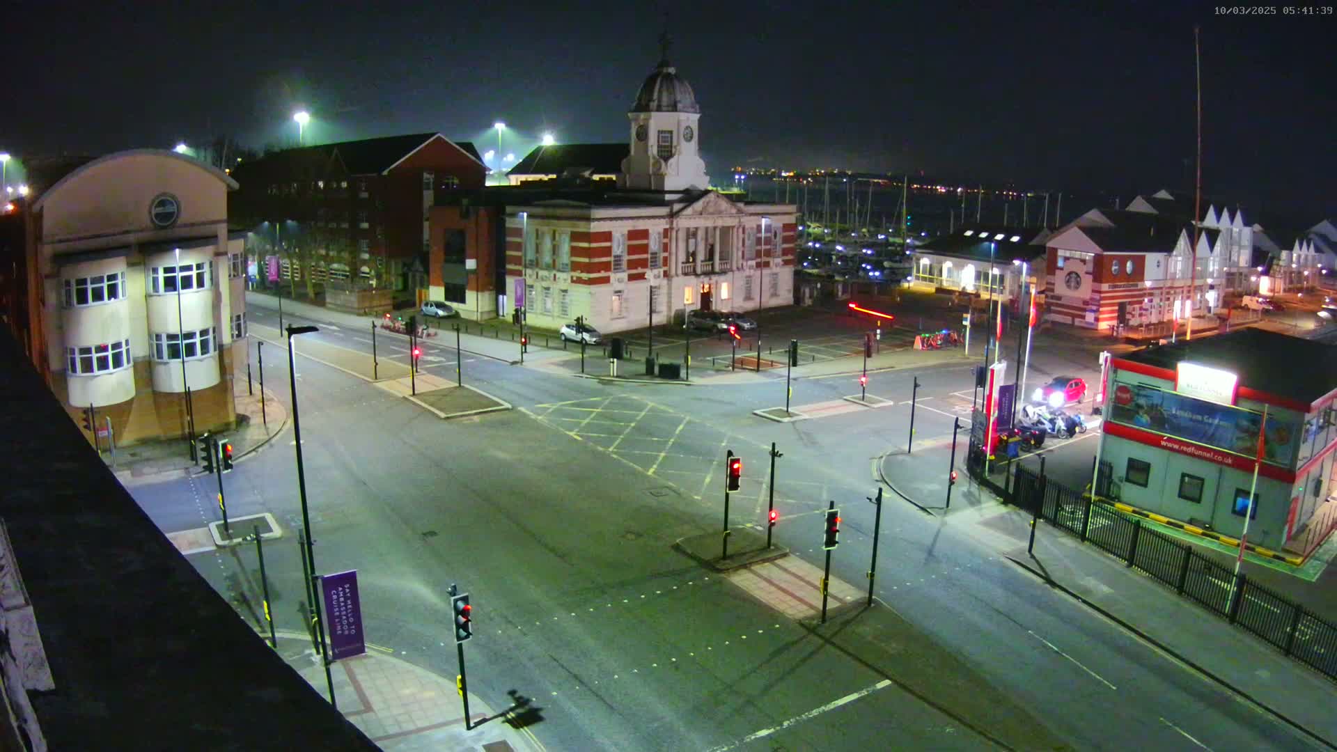 A nighttime, high-angle view of a town square with a large brick building, several smaller buildings, and a marina in the background, illuminated by streetlights.