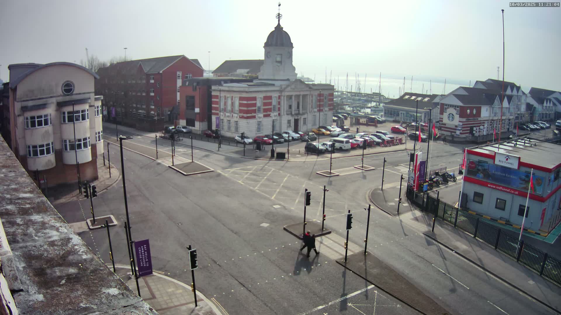 A high-angle view of a town square with a large brick building, surrounding buildings, parked cars, a pedestrian crossing, and a body of water with boats in the distance under a hazy, sunny sky.