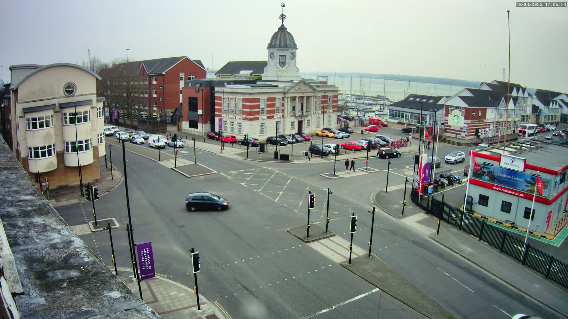 A busy intersection with several buildings, including a large clock tower building,  is seen on an overcast day, with a body of water visible in the background.