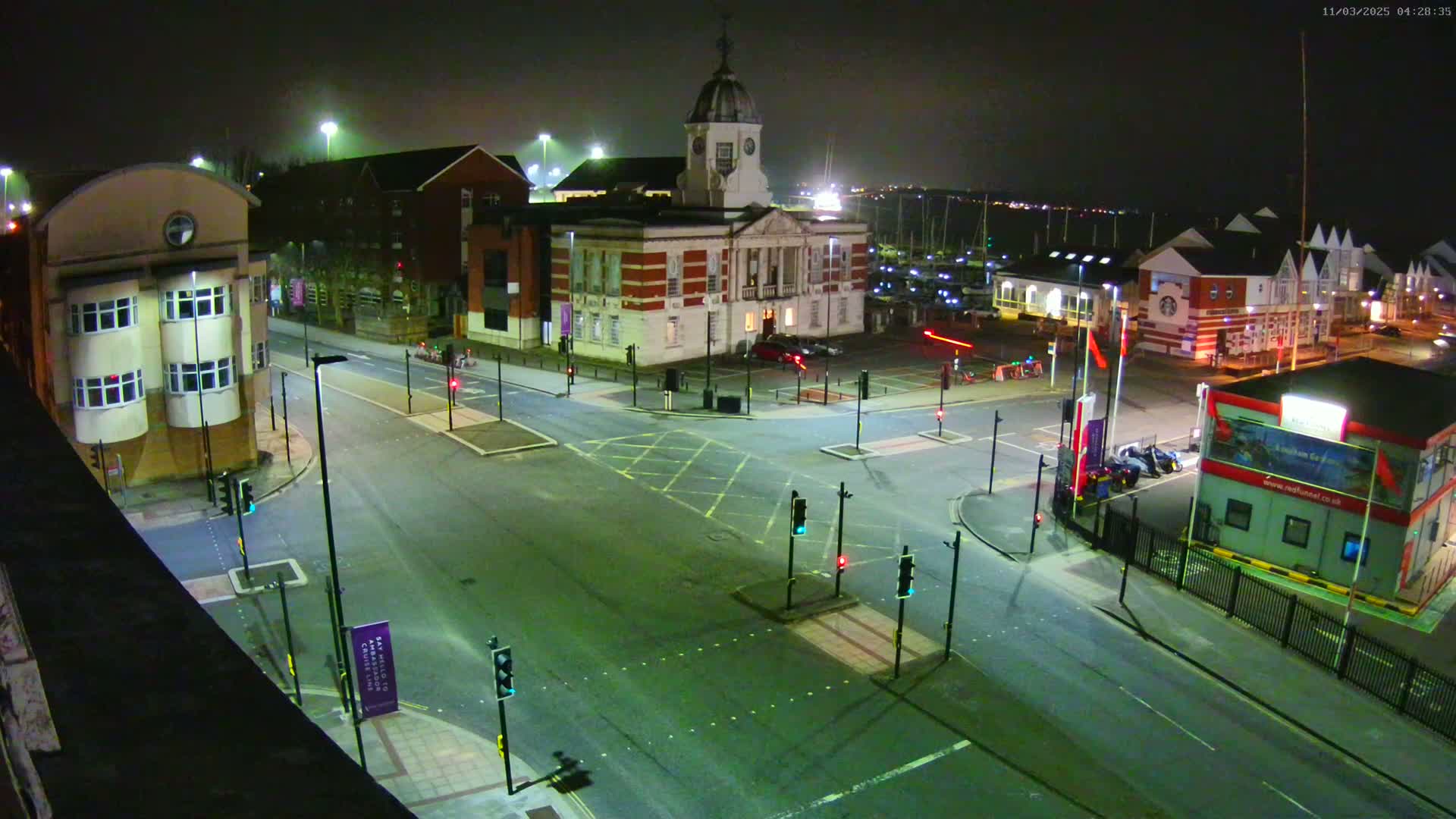 A nighttime aerial view of a mostly empty intersection with traffic lights, several buildings, and a marina in the background under a dark, clear sky.