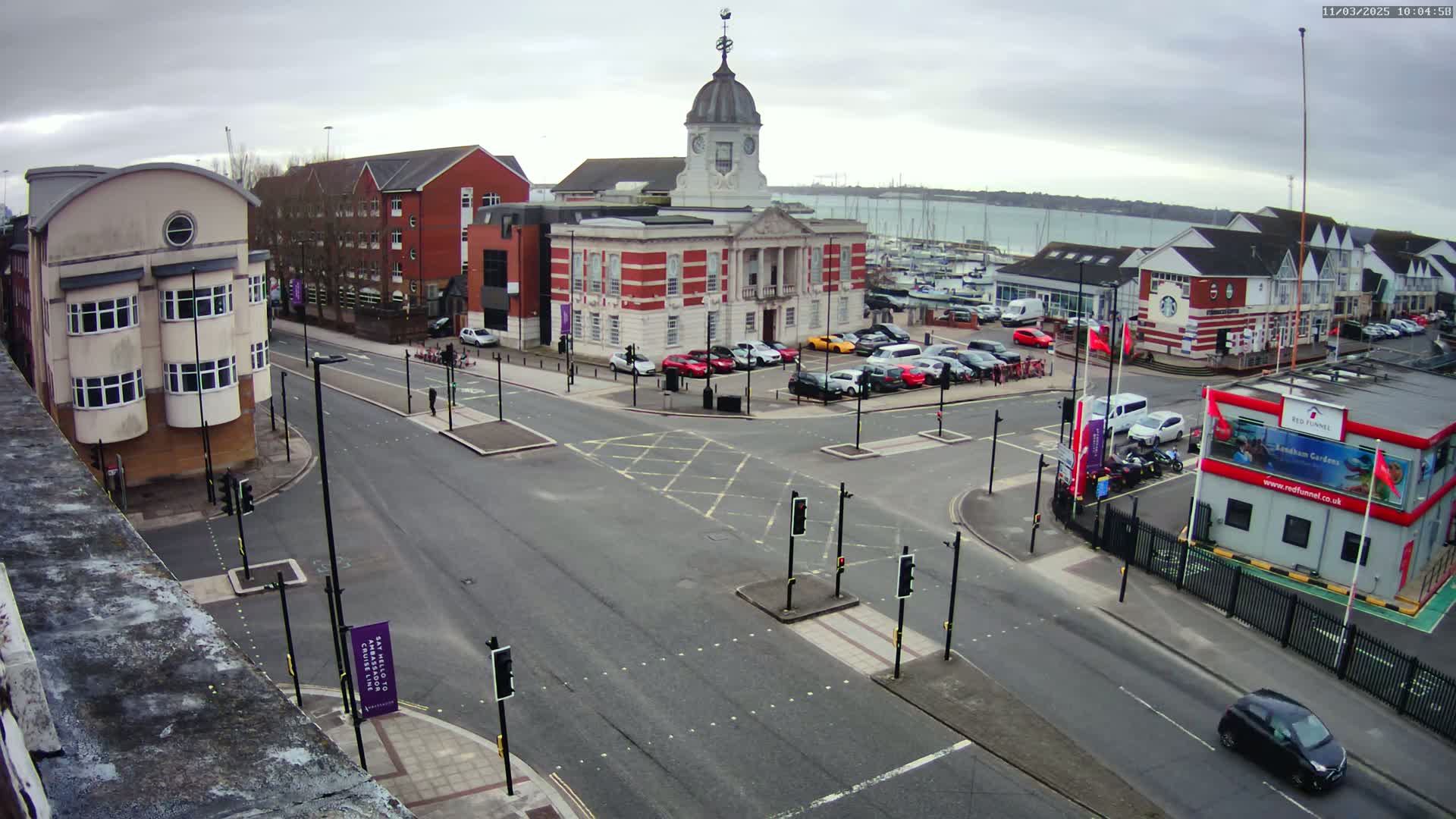 A town square with a brick building and clock tower is viewed from above on an overcast day, with a crossroads and parked cars surrounding it.