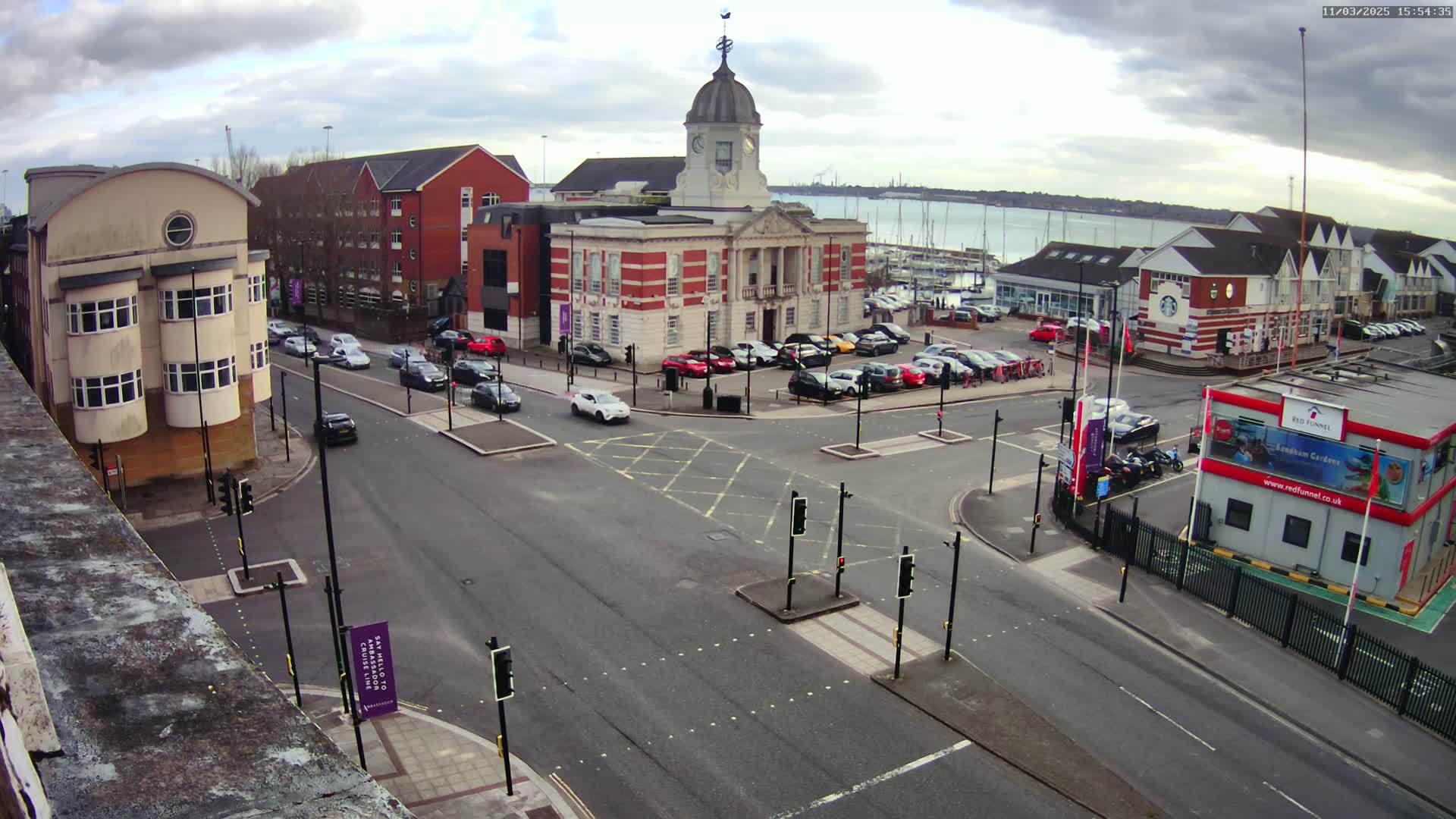 A street intersection with several cars, a large brick building with a clock tower, and a marina with boats under a cloudy sky.
