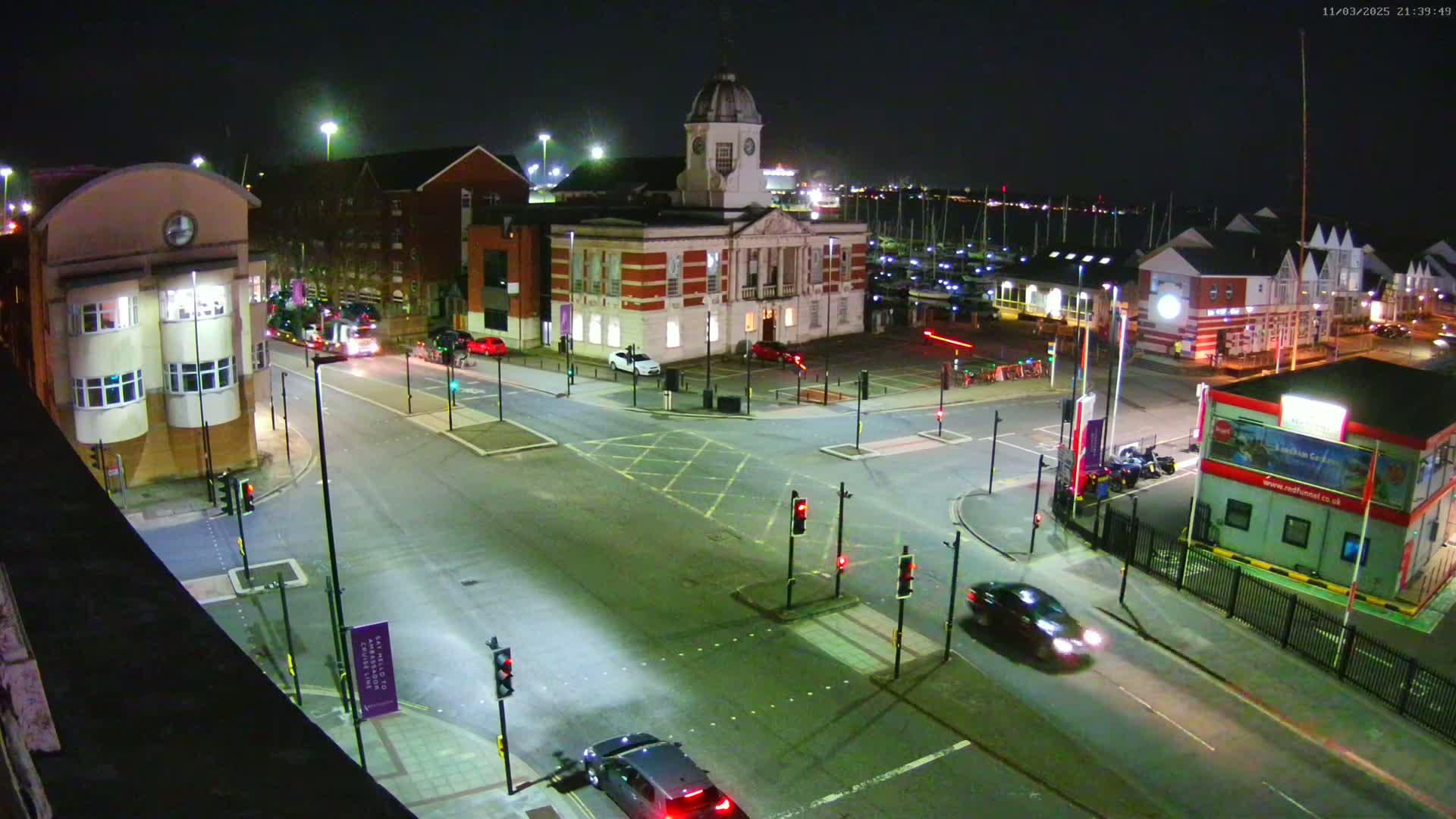 A nighttime aerial view of a town square with a large brick building, several cars, and traffic signals at a crossroads, under clear skies.