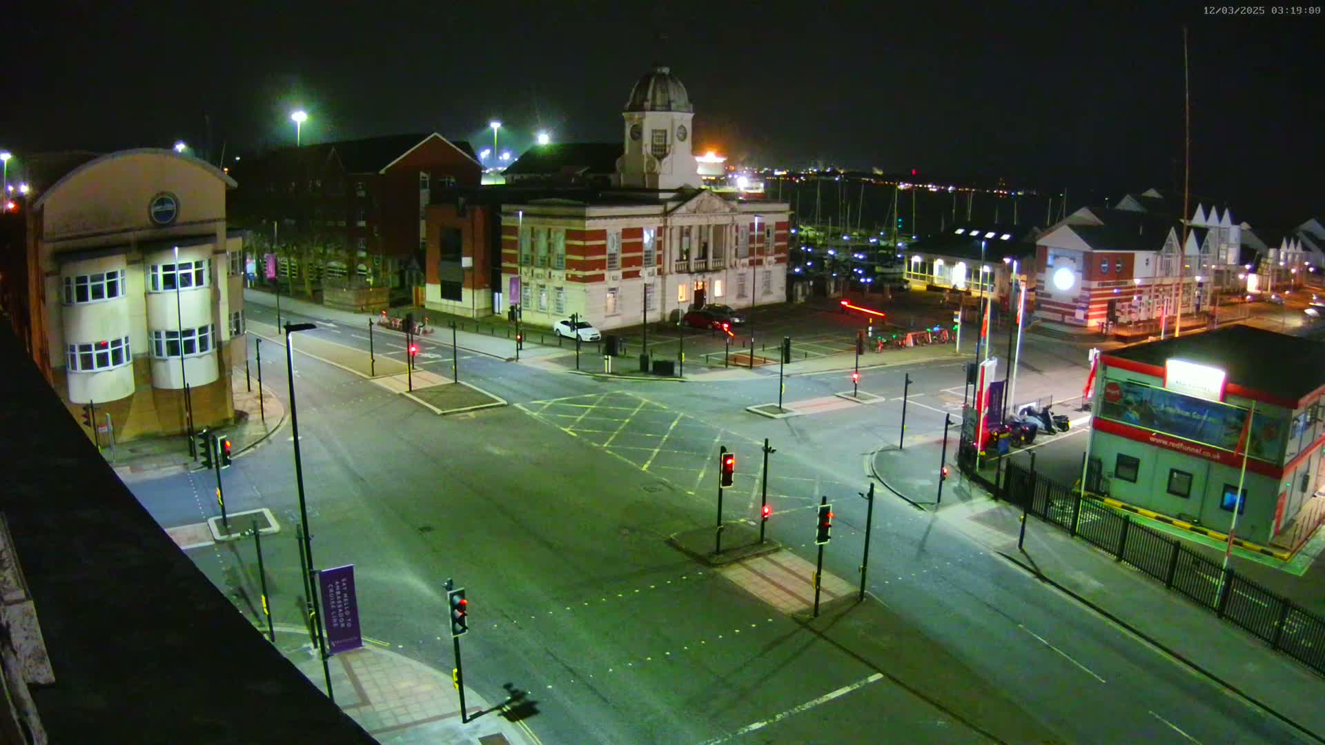 A nighttime aerial view of a mostly empty intersection in front of several buildings, including one with a clock tower, near a marina.