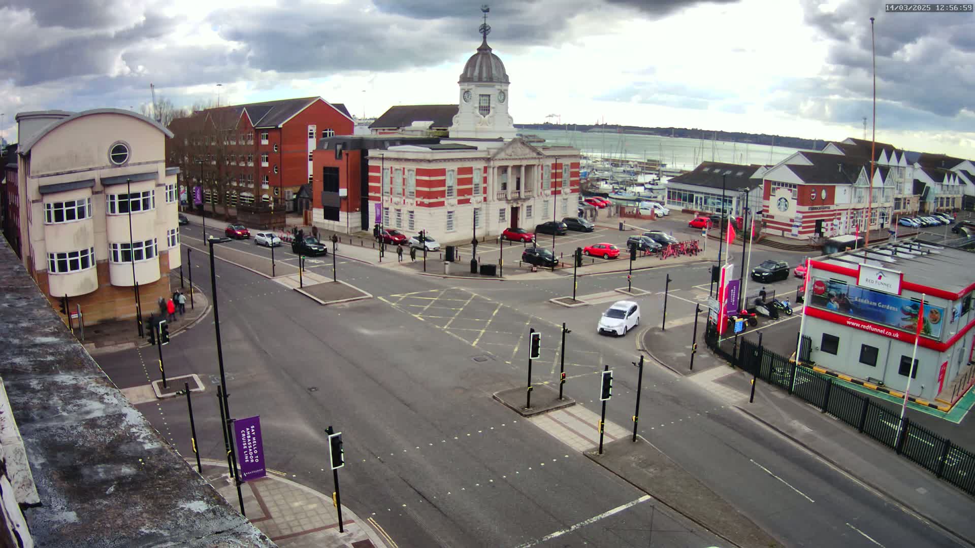 A street intersection with a large brick building, other buildings, and parked cars, under a cloudy sky, overlooks a marina with boats in the distance.