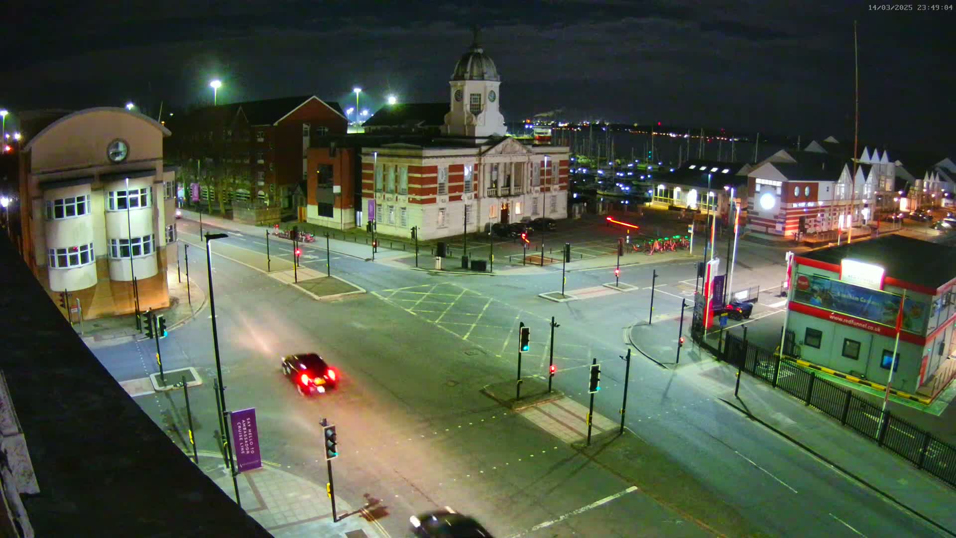 A nighttime aerial view of a town square with a large clock tower building, several other buildings, roads, traffic lights, and a few cars.