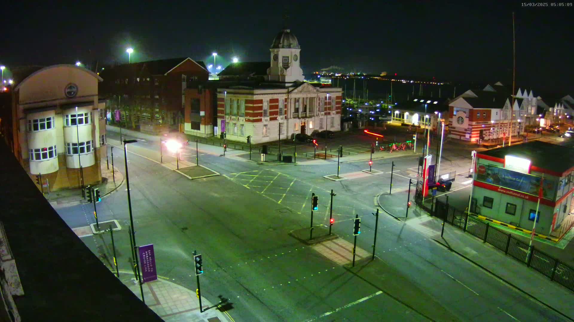 An empty nighttime intersection with traffic signals and buildings surrounding a harbor visible in the background under clear skies.