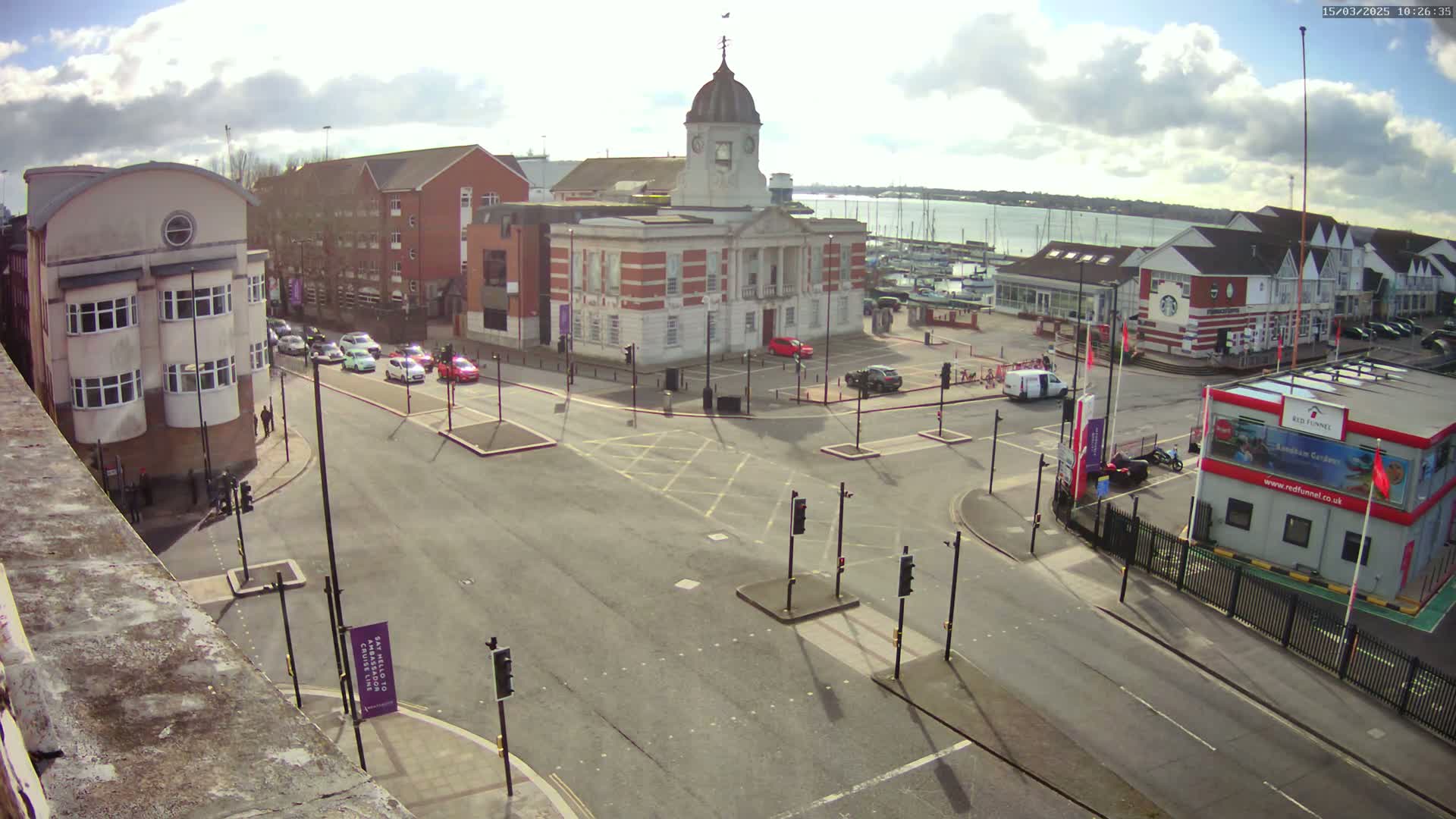 A mostly sunny day reveals a town square with a brick building and clock tower at its center, surrounded by other buildings, roads with cars, and a harbor with boats in the distance.