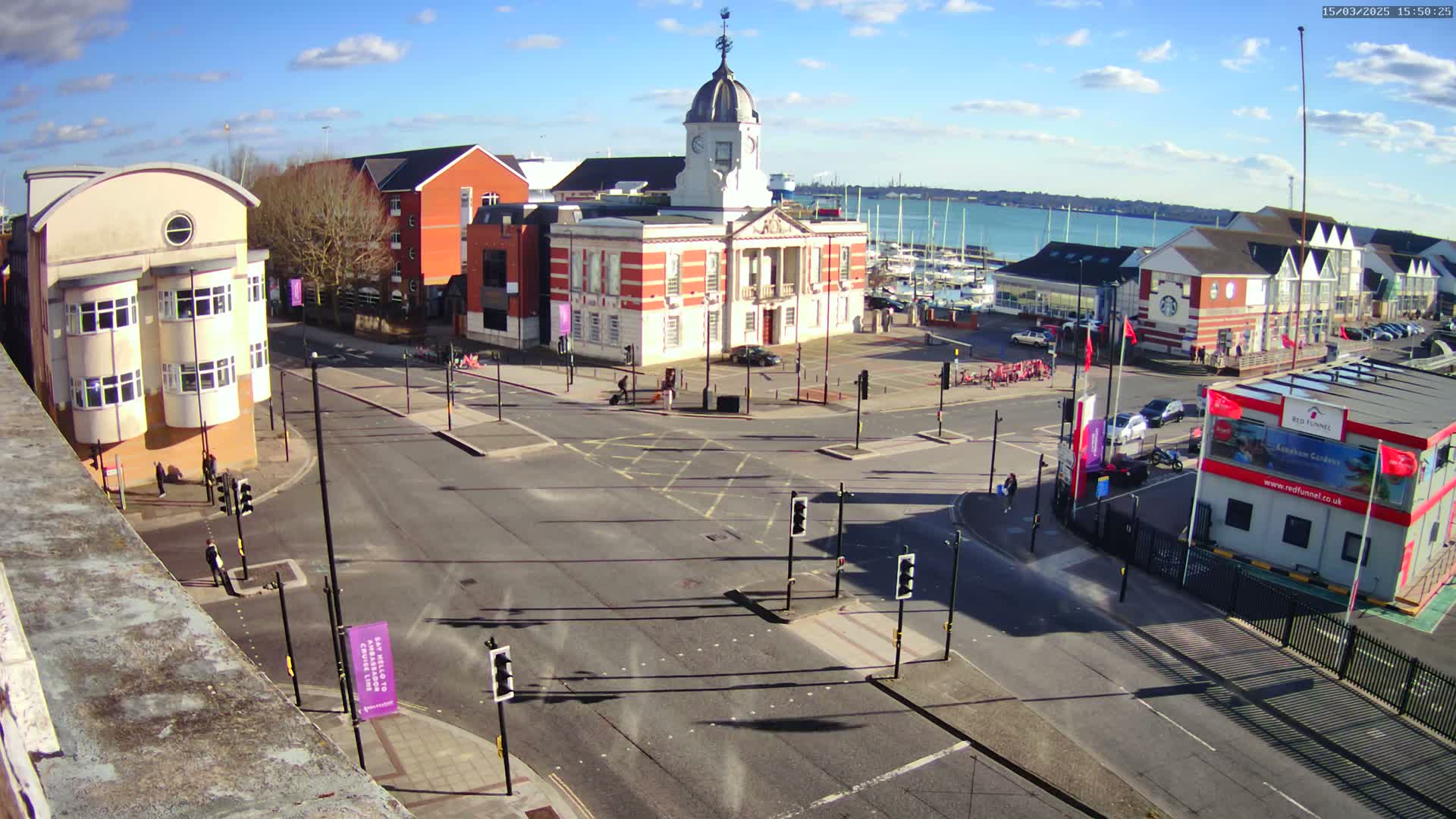 A sunny day shows a town square with a large brick building at its center, surrounded by other buildings and a harbor with boats visible in the distance.