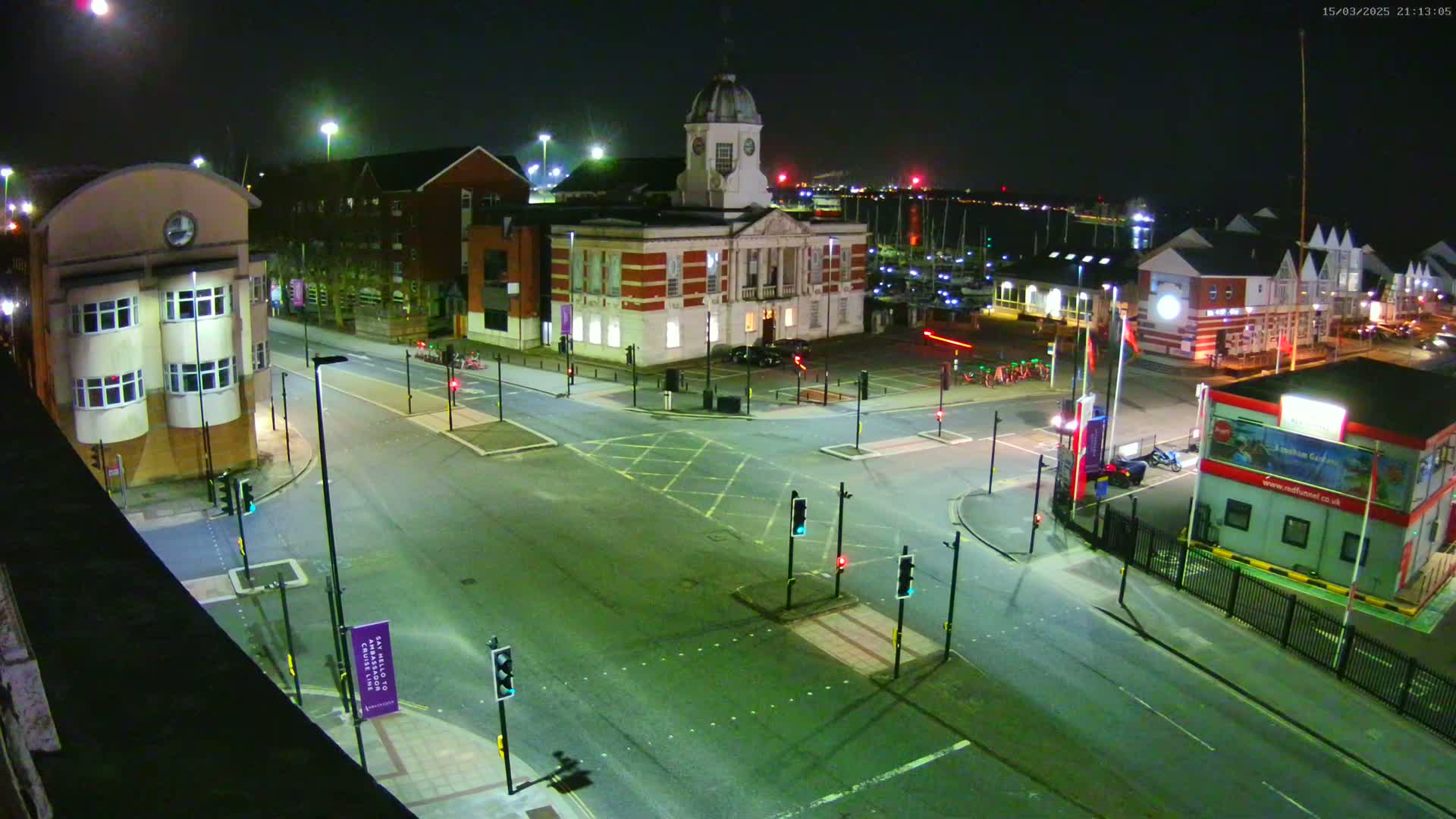 An empty, well-lit intersection at night, surrounded by buildings including a clock tower and marina, under a clear, dark sky.