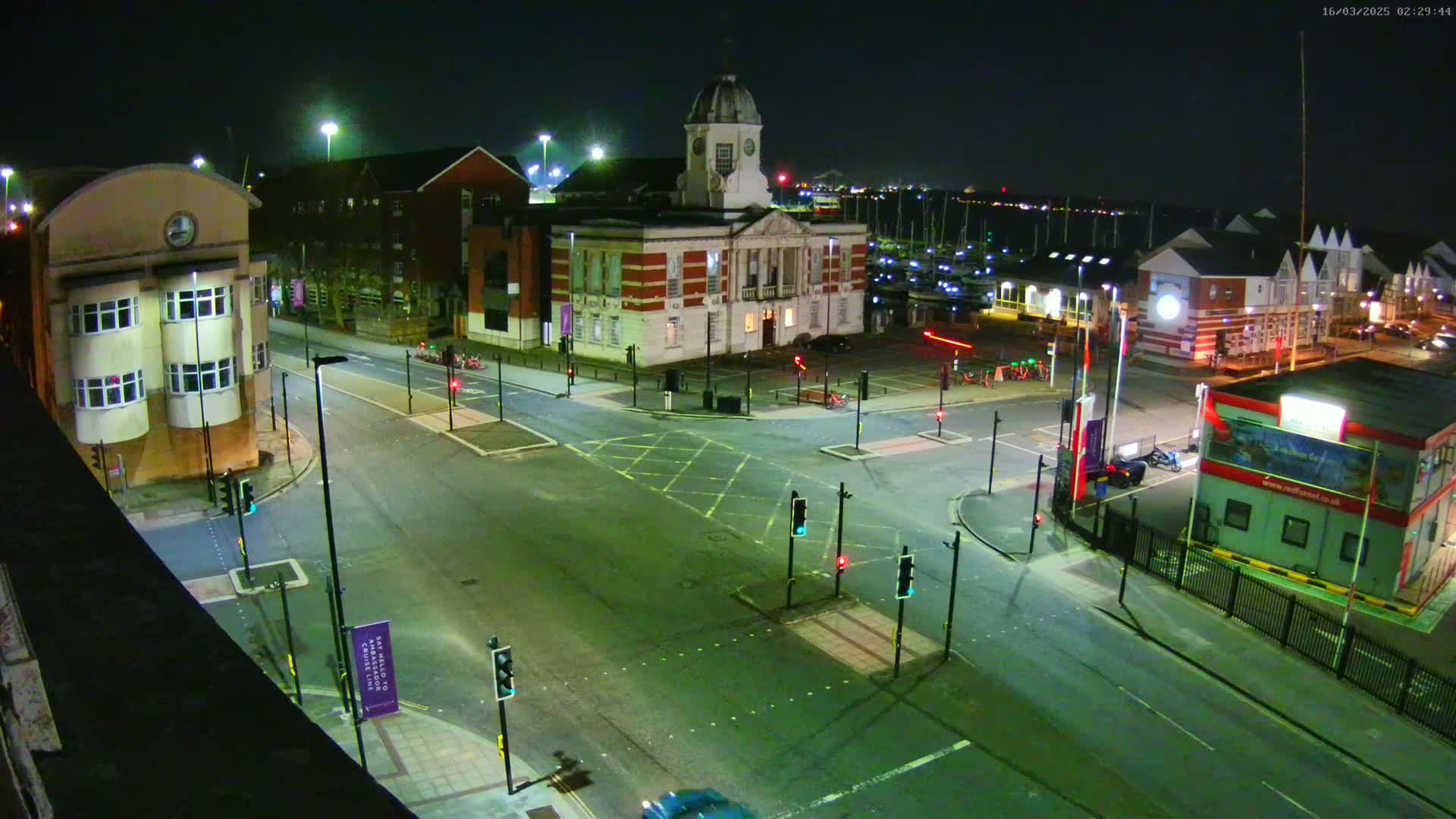 A nighttime, high-angle view of a town square with several buildings, including a large brick building with a clock tower, surrounded by roads and traffic signals.
