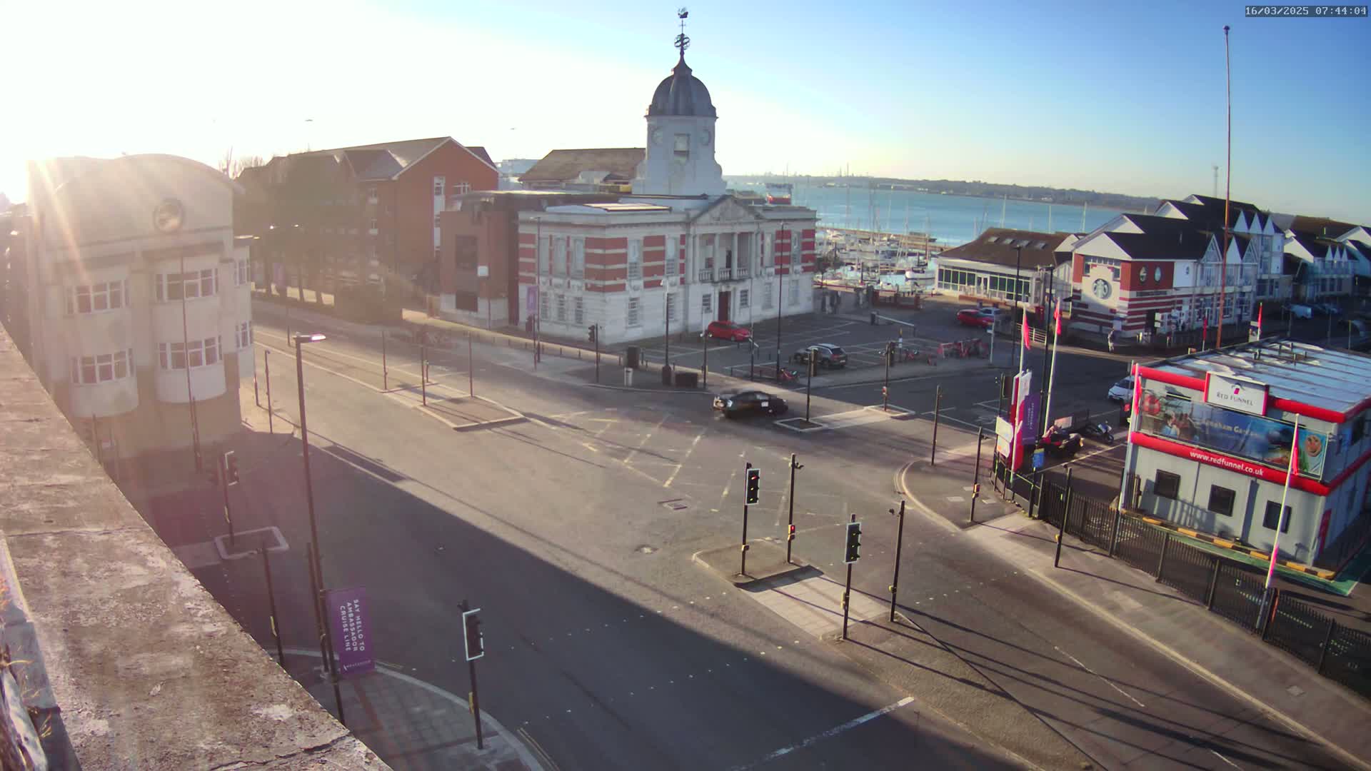 A sunny day reveals a harbor town square with a large red brick building, several other buildings, cars, and a marina in the background.