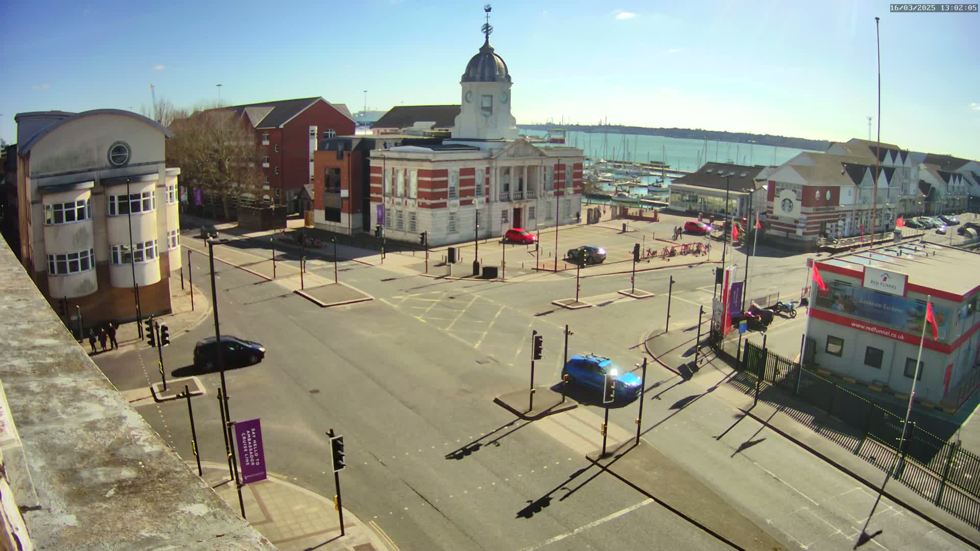 A sunny day overlooking a town square with a brick building, several cars, and a marina in the background.