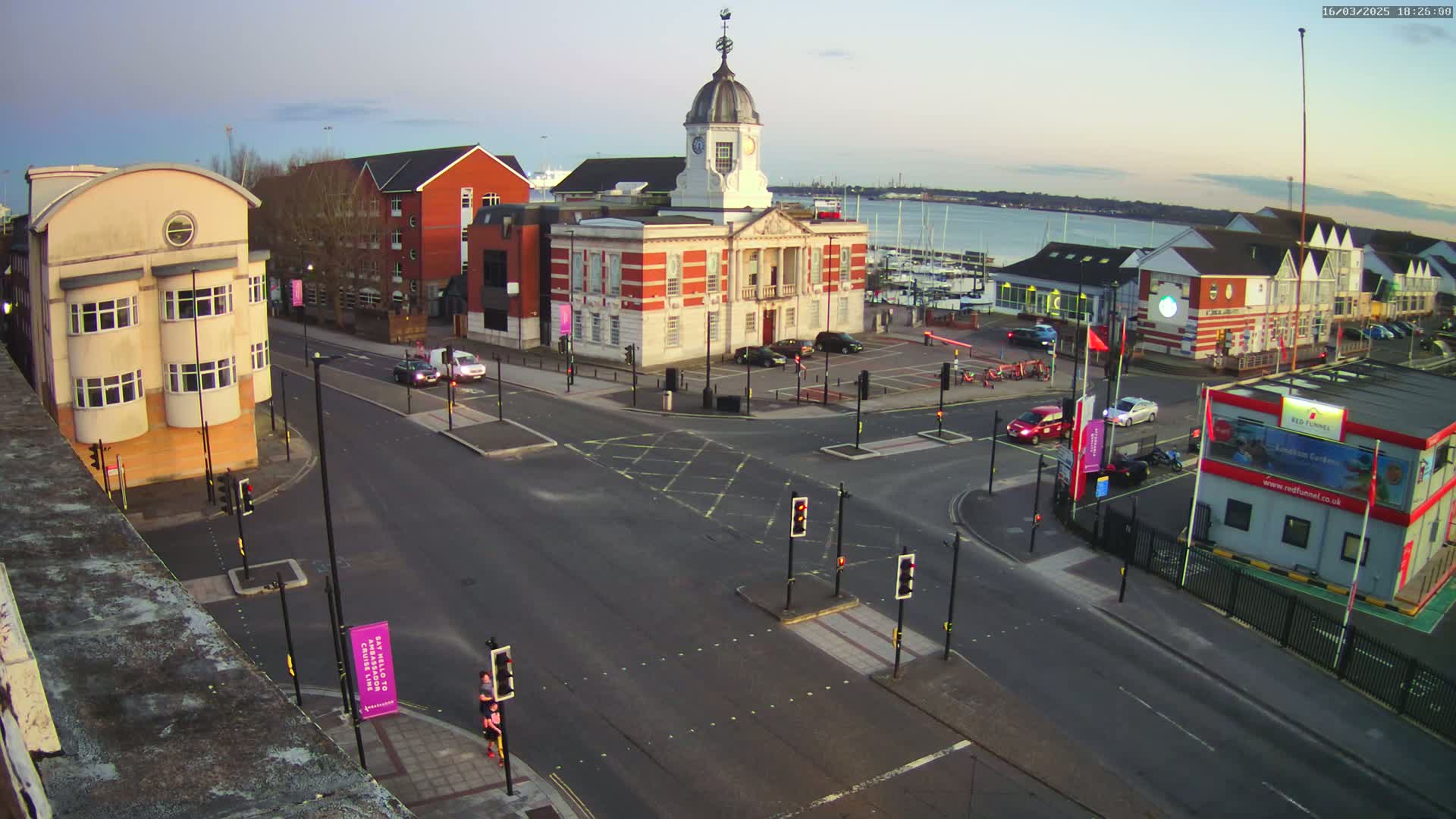 A high-angle, twilight view of a town square with a prominent clock tower building, surrounding buildings, a roadway intersection with traffic signals, and a body of water visible in the distance.