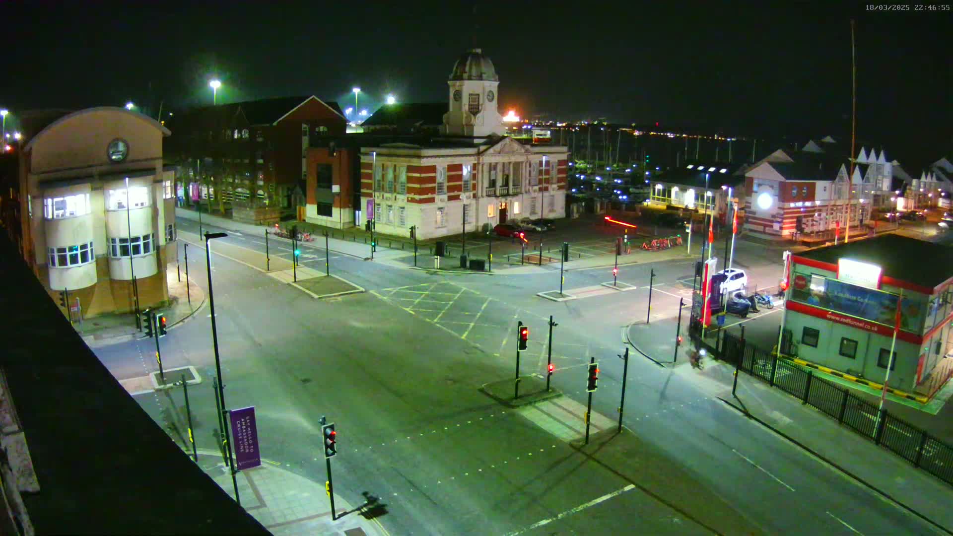 A nighttime, high-angle view of a town square with a large brick building at its center, surrounded by roads with traffic lights and parked cars.