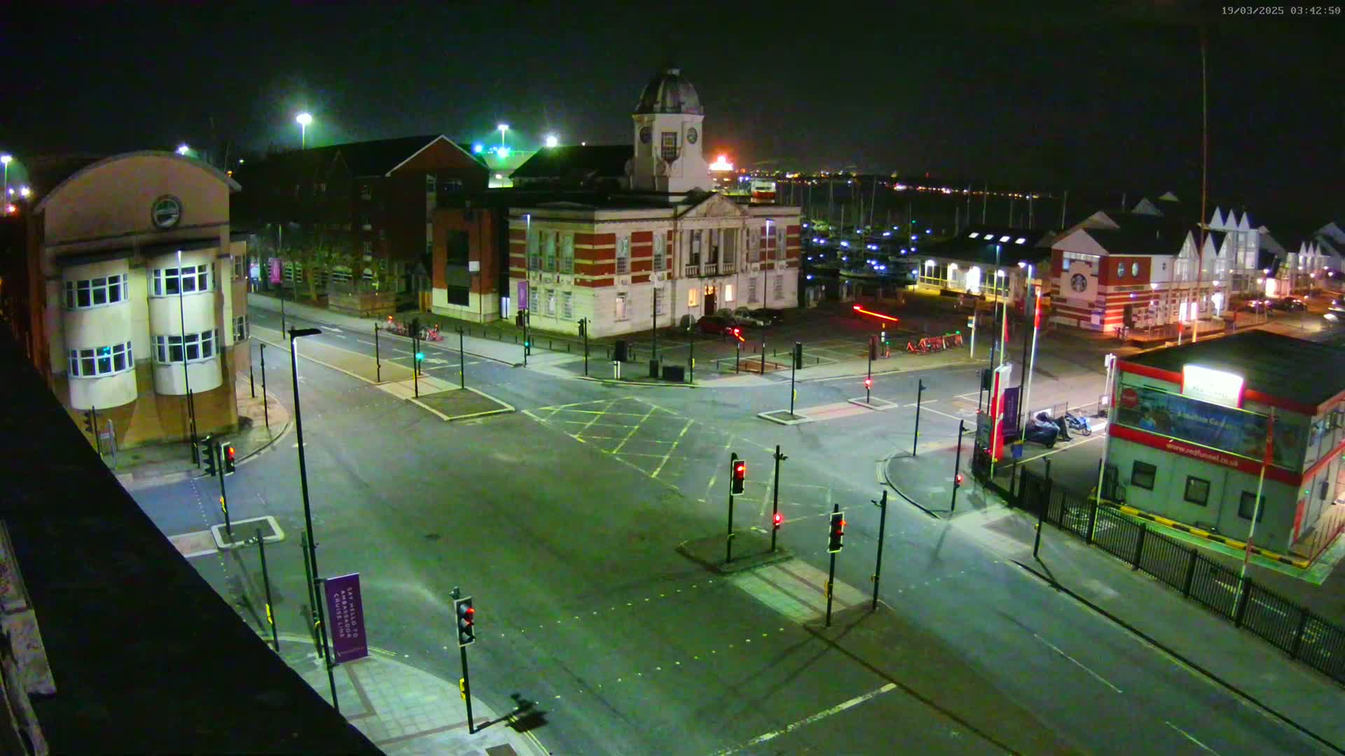 An empty nighttime intersection with traffic lights, surrounded by buildings including a large clock tower building and smaller shops near a body of water.