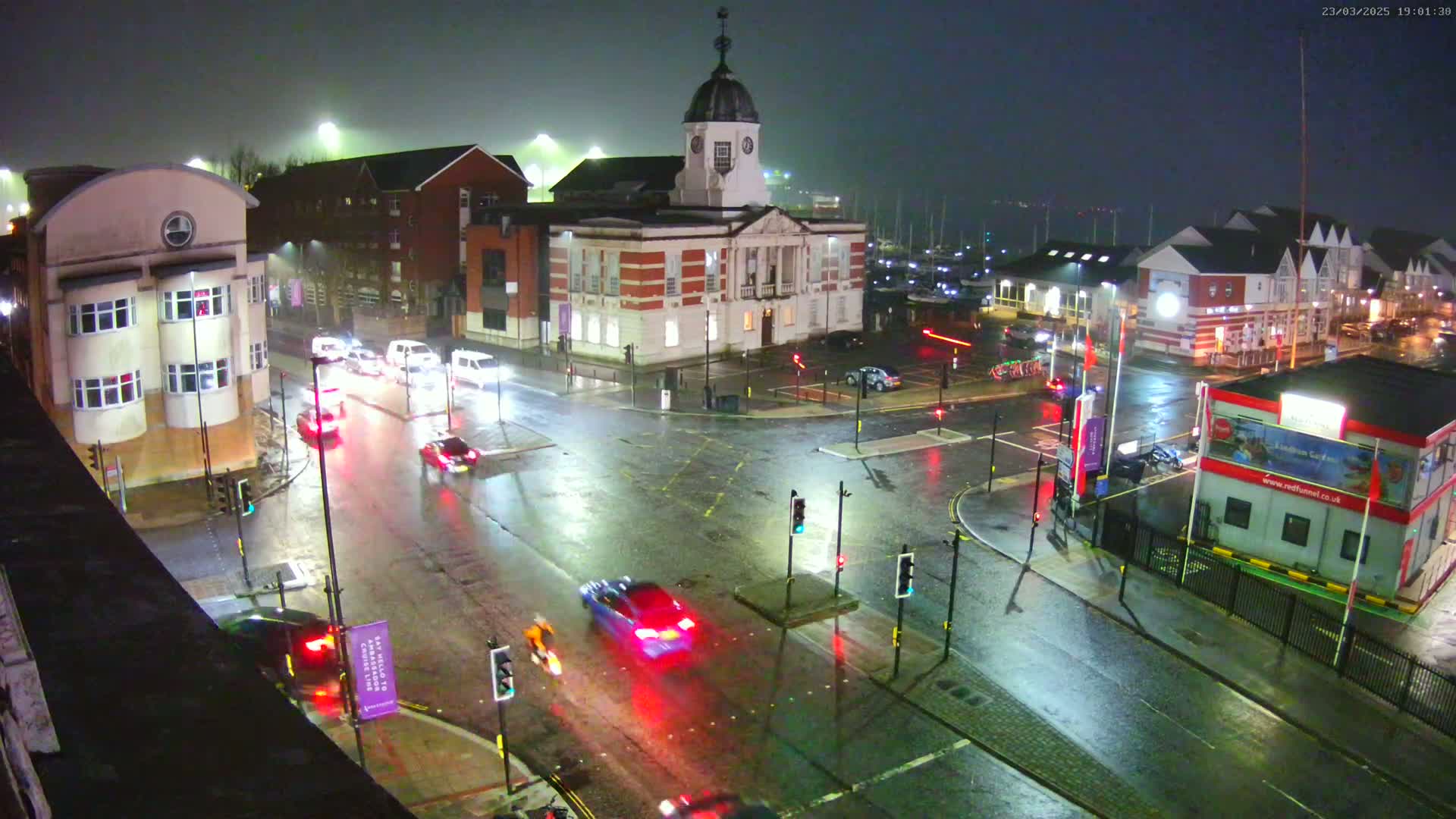 A rain-slicked intersection at night, showing several cars and buildings, including a prominent clock tower building, under nighttime, rainy conditions.
