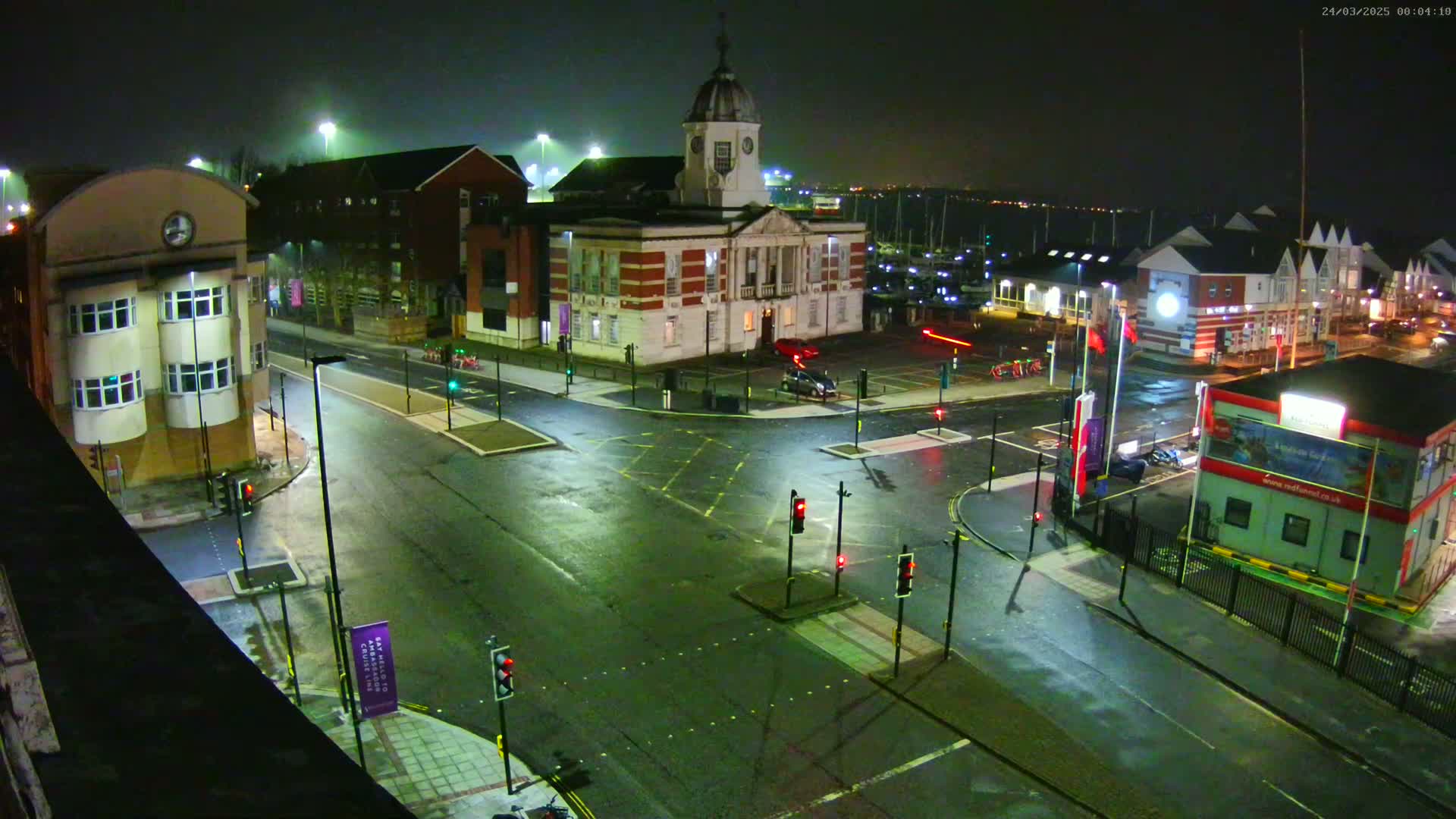 A wet, nighttime street intersection showing several buildings, including a large brick building with a cupola, and a marina in the background.