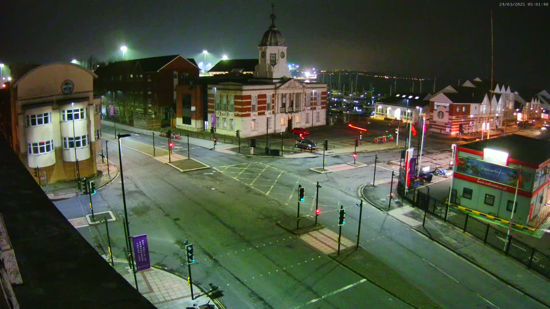 A nighttime, aerial view of a street intersection near a waterfront, featuring several buildings, including a large brick building with a cupola, under mostly clear skies.