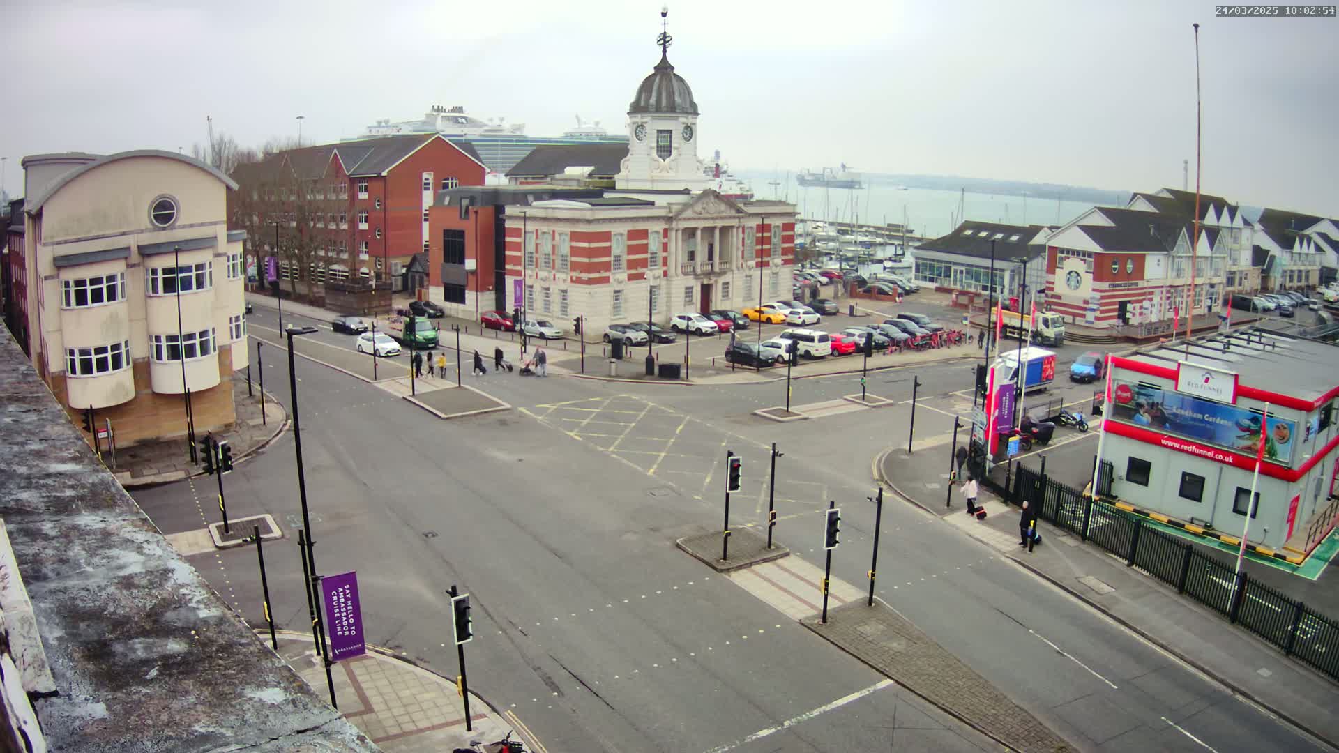 An overcast day reveals a city intersection with a brick building and clock tower at its center, surrounded by parked cars, pedestrians, and buildings, all overlooking a marina and a body of water beyond.