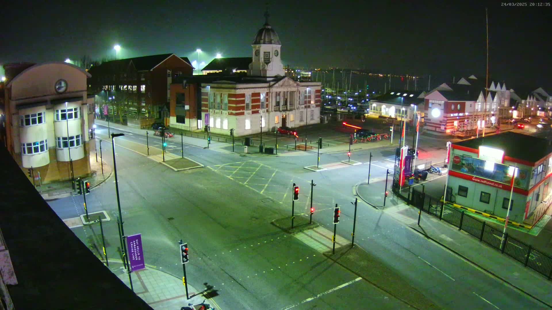 A nighttime, high-angle view of a town square with a clock tower building and several other buildings, roads, and traffic lights, with a body of water visible in the background.