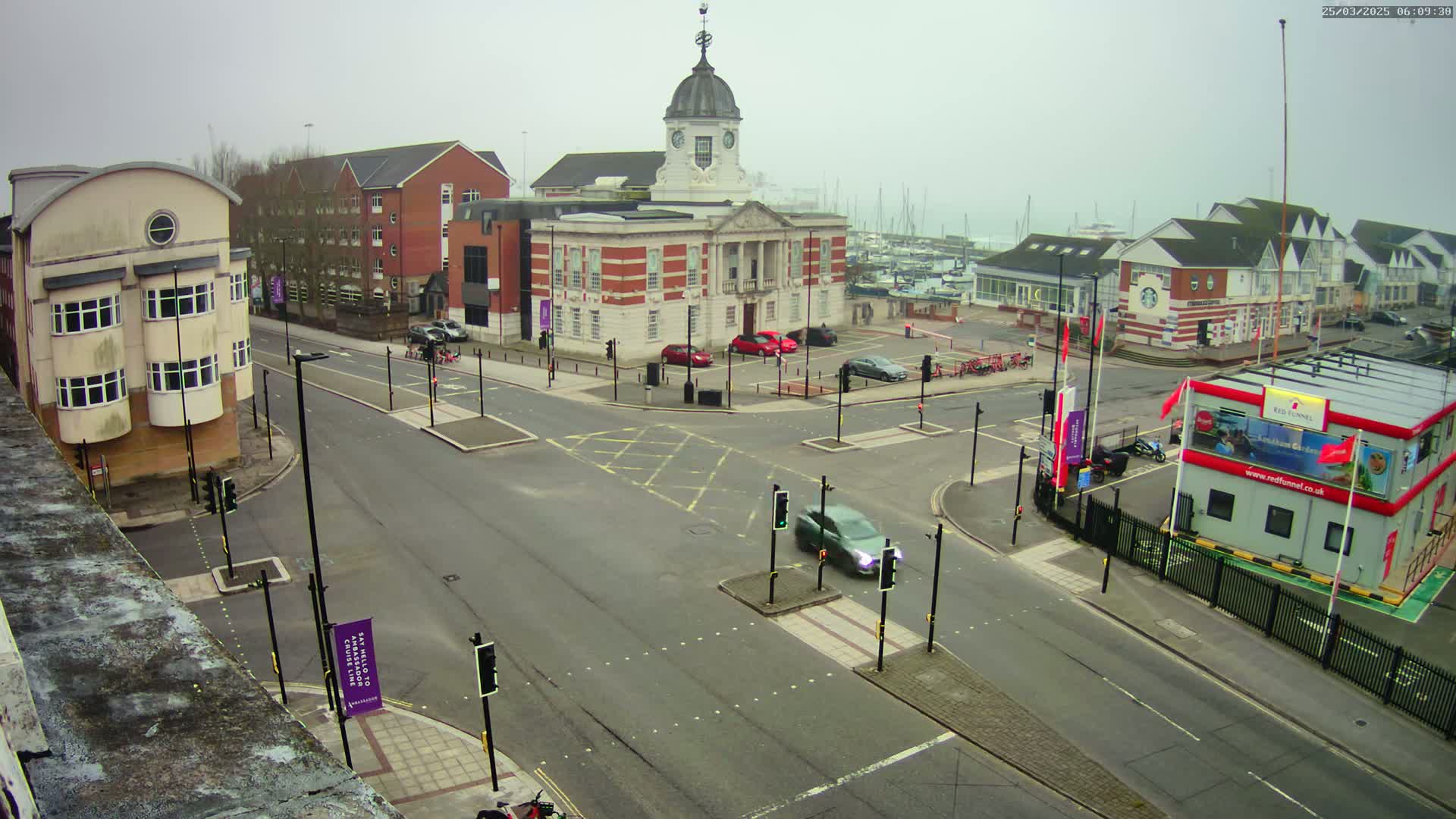 A high-angle view of a city intersection on an overcast day, showing several buildings, parked cars, and a few vehicles in motion.