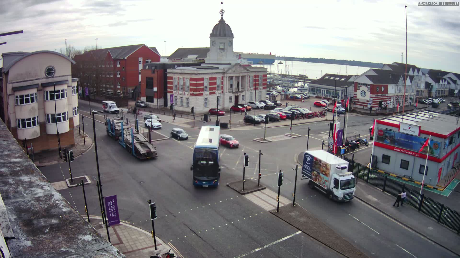 A high-angle view of a town square with several vehicles at an intersection on an overcast day, featuring a brick building with a dome in the center and a body of water in the background.