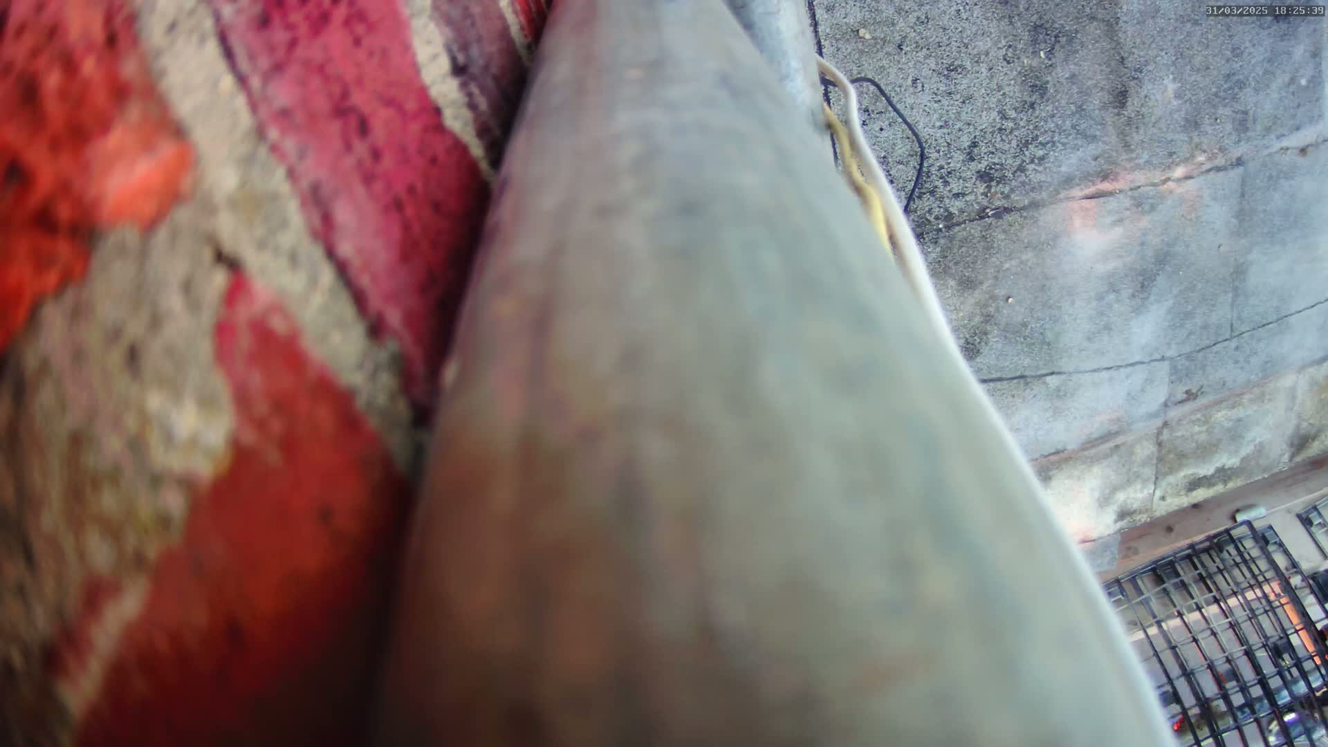 A weathered metal pipe is positioned against a red brick wall, with a grey concrete surface visible in the background.