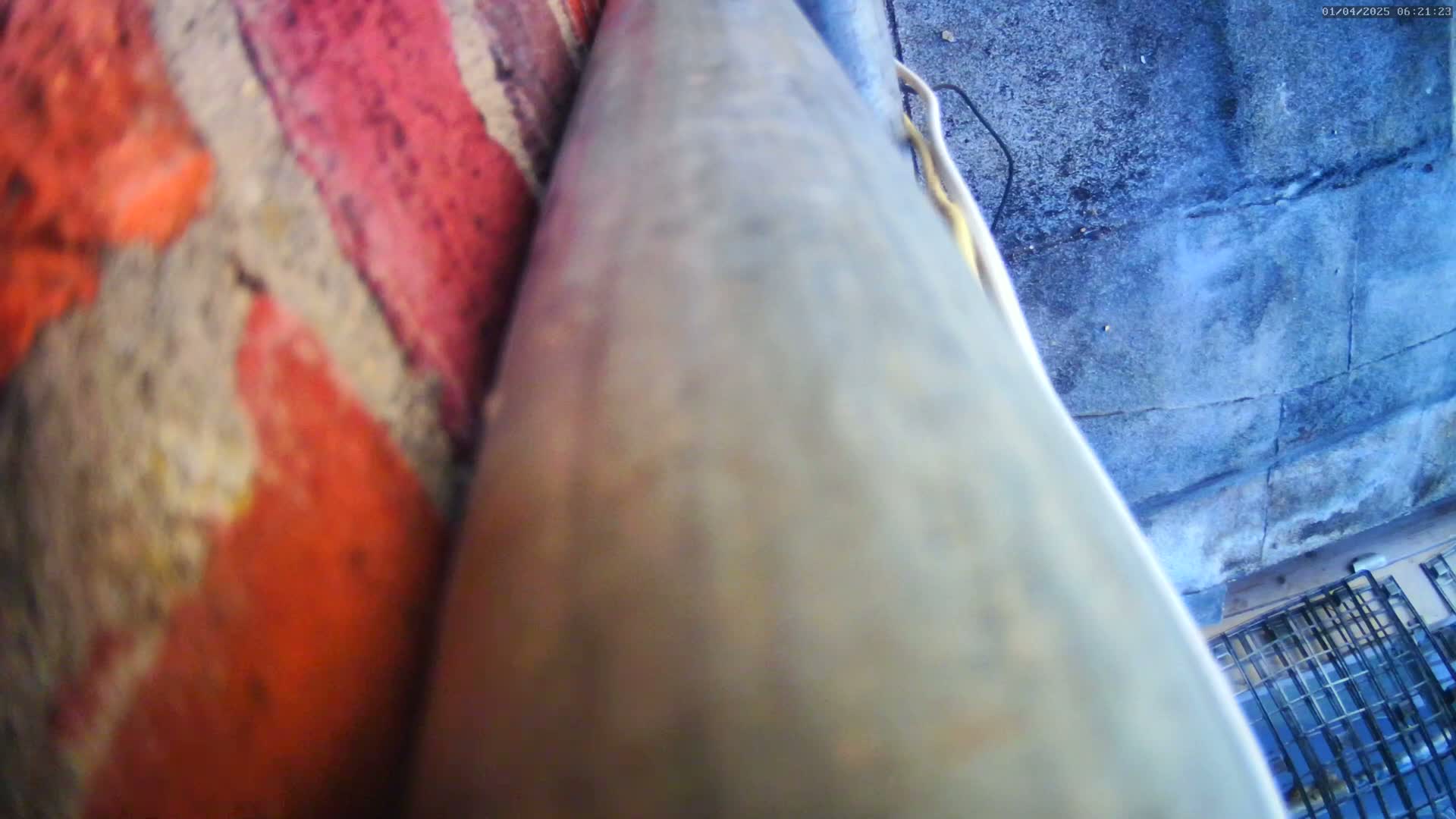A close-up view shows a weathered gray surface next to a reddish-brown brick wall, with a large, light-colored metallic object angled between them.