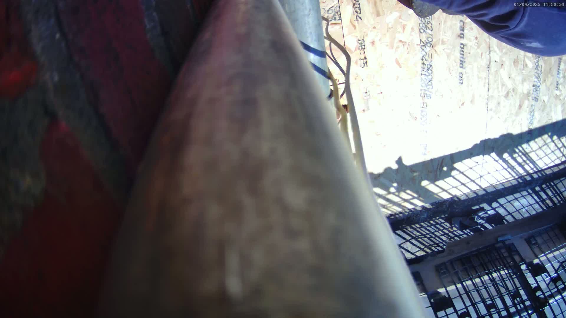 A close-up, low-angle view shows a dirty, metallic object angled towards the viewer, partially obscuring a view of a light-colored wooden surface and a metal grating in bright, sunny conditions.