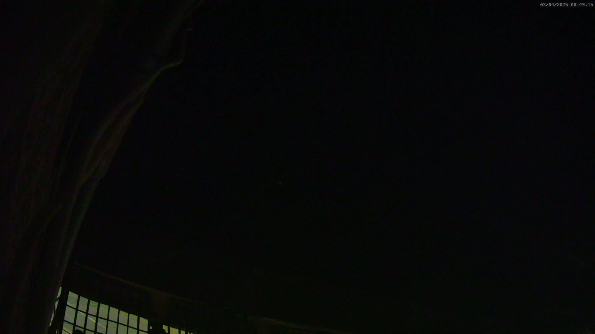 A dark, nighttime scene shows a portion of a building with a window and a large, dark object, possibly a wall or curtain, extending into the frame.