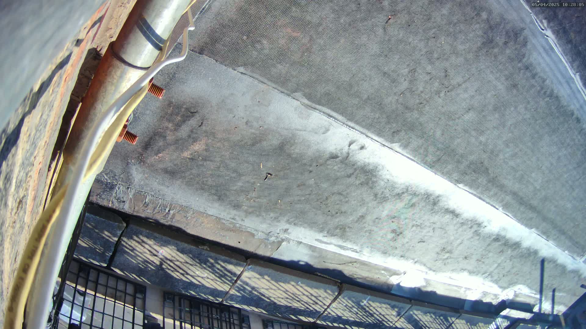 A high-angle view of a weathered gray rooftop on a sunny day, showing a metallic pipe and fastening hardware attached to it.